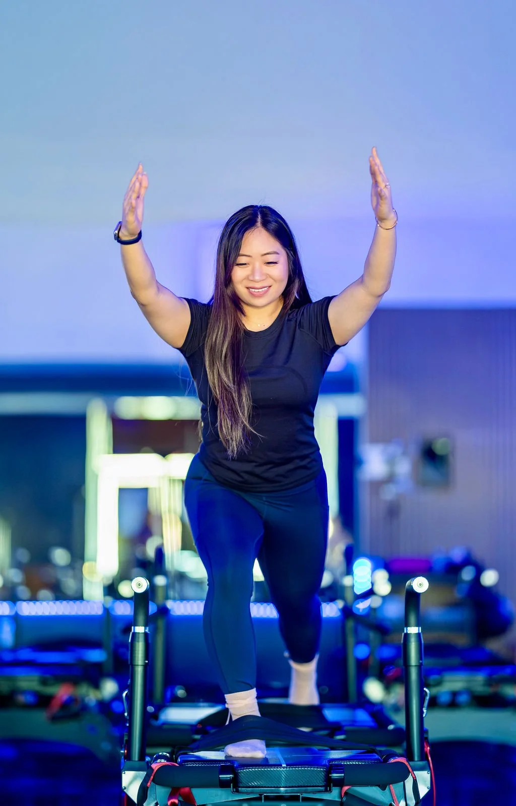 Woman stands in a lunge on a Maven reformer with both arms raised, smiling, in a Persona Pilates studio