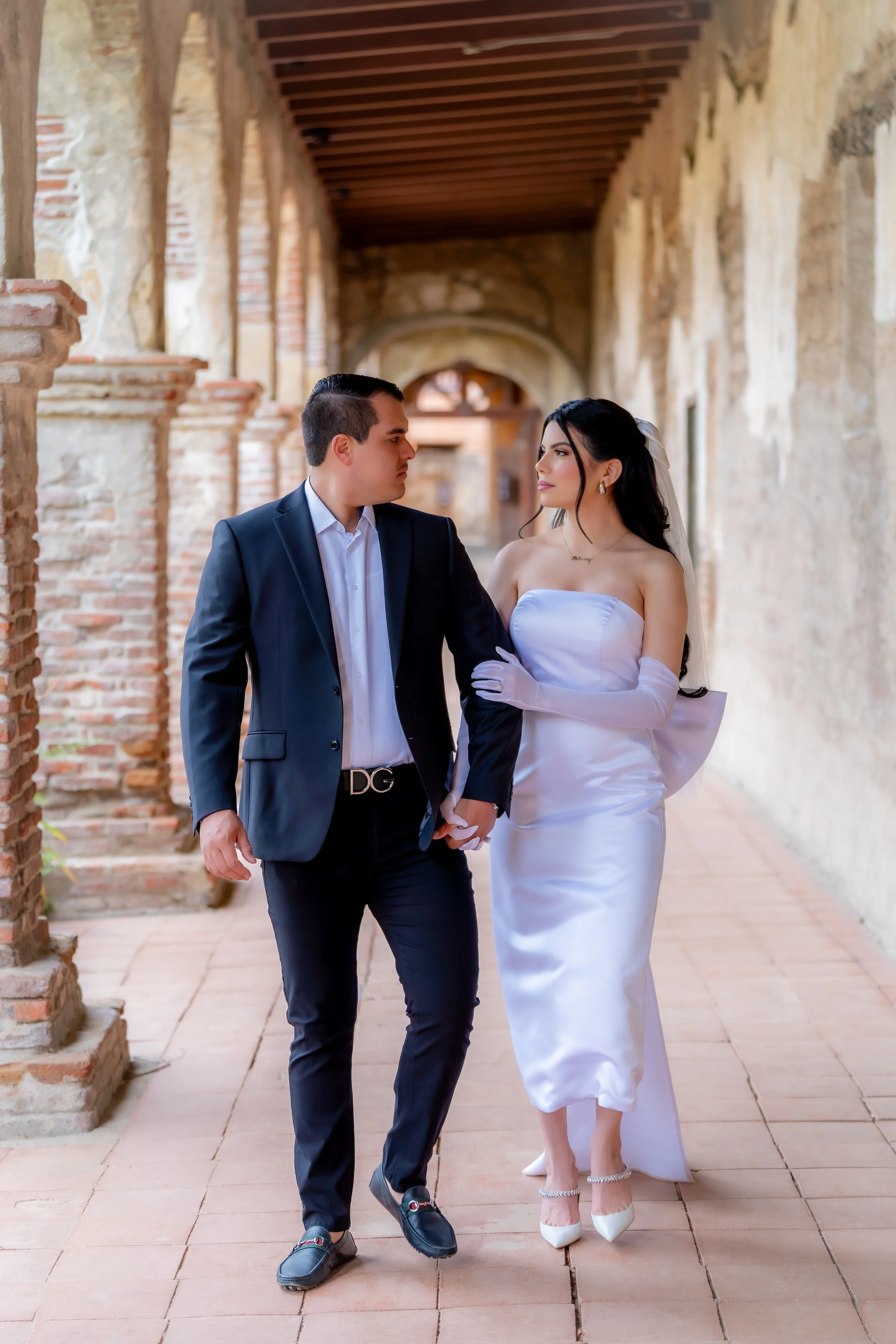 A couple dressed in wedding attire walking hand in hand in a rustic, brick archway corridor.