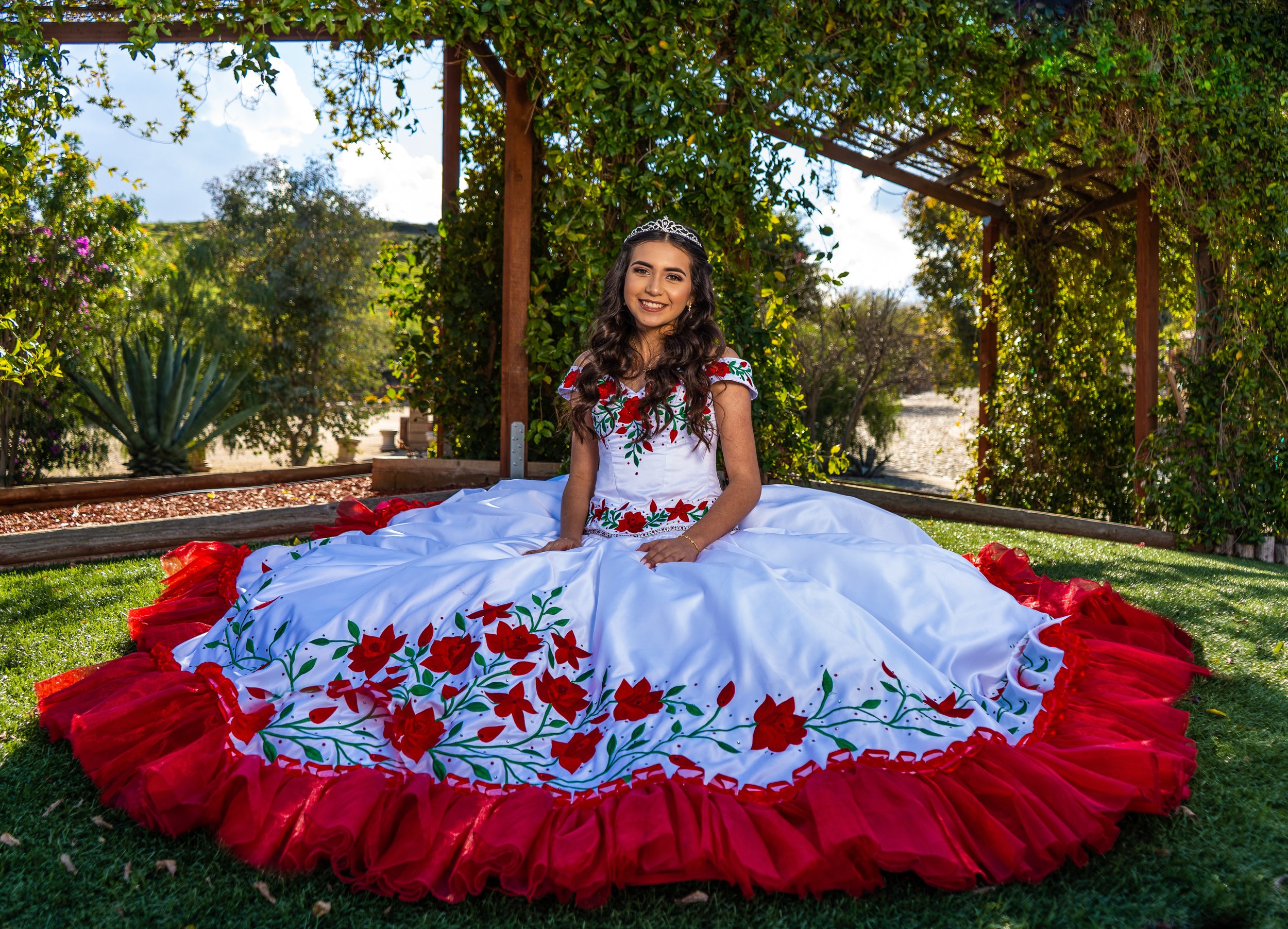 A girl wearing a white dress with red and green floral embroidery and red tulle trim, sitting outdoors on green grass under a wooden pergola with green foliage. She is smiling and wearing a tiara.
