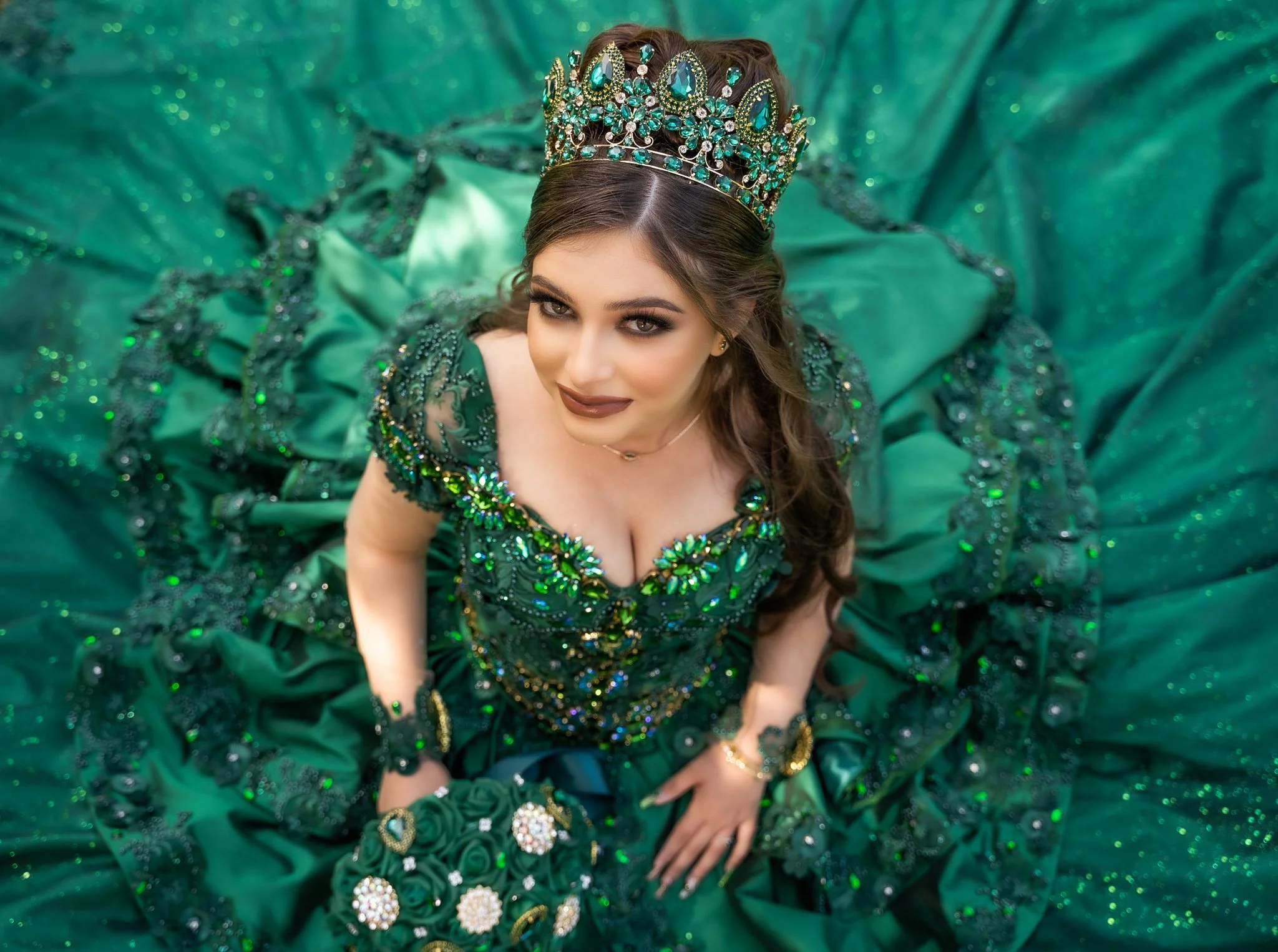 A woman wearing a green, embellished gown and a jeweled crown, looking up at the camera on a matching green satin backdrop.