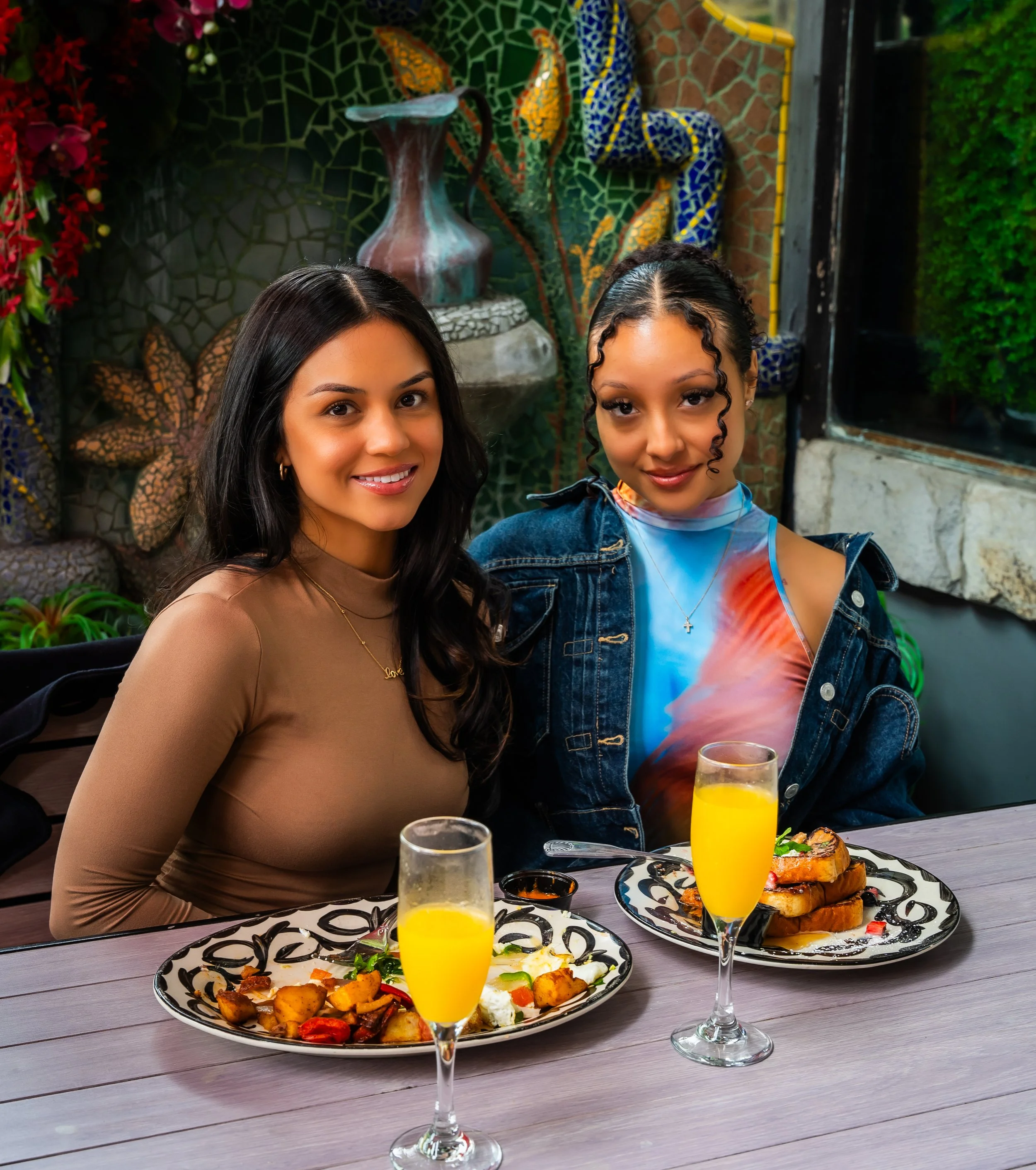 Two women sitting at a table with breakfast, each with a plate of food and a glass of orange juice, smiling for the camera in a decorative restaurant setting.