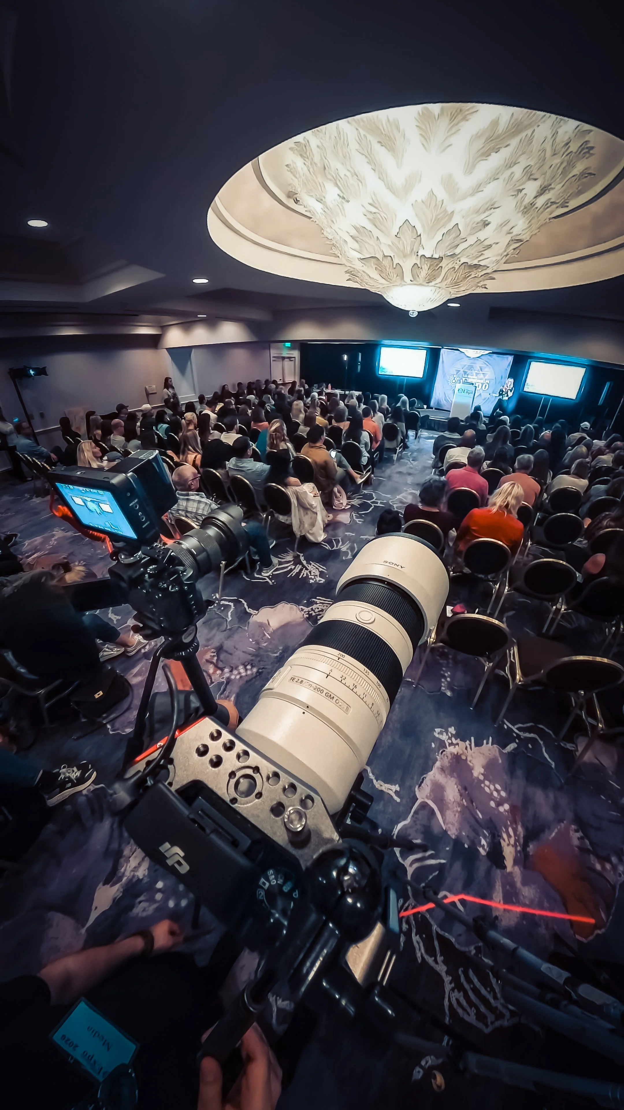 A conference or seminar room filled with attendees watching a presentation. showing a 2 camera setup for a wide and closed up, The photo is taken from a mounted camera with a large telephoto lens, pointing towards the audience.