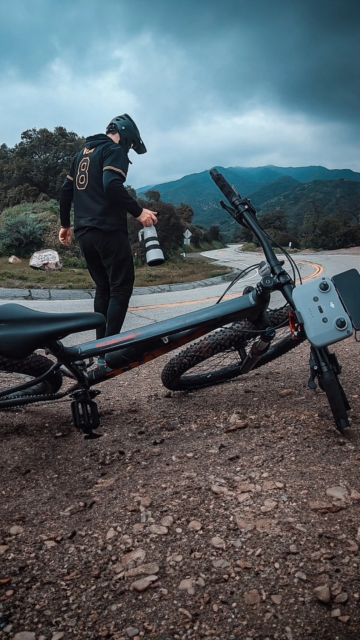 A rider, shooter on a mountain road, holding a large camera lens, with an mtb bike lying on the gravel next to them with a drone control attached. Scenic mountains and cloudy sky in the background.