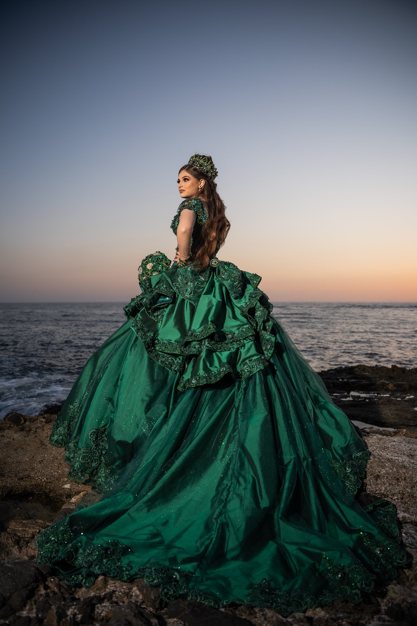 A woman in an elaborate green gown standing on rocks by the ocean at sunset.
