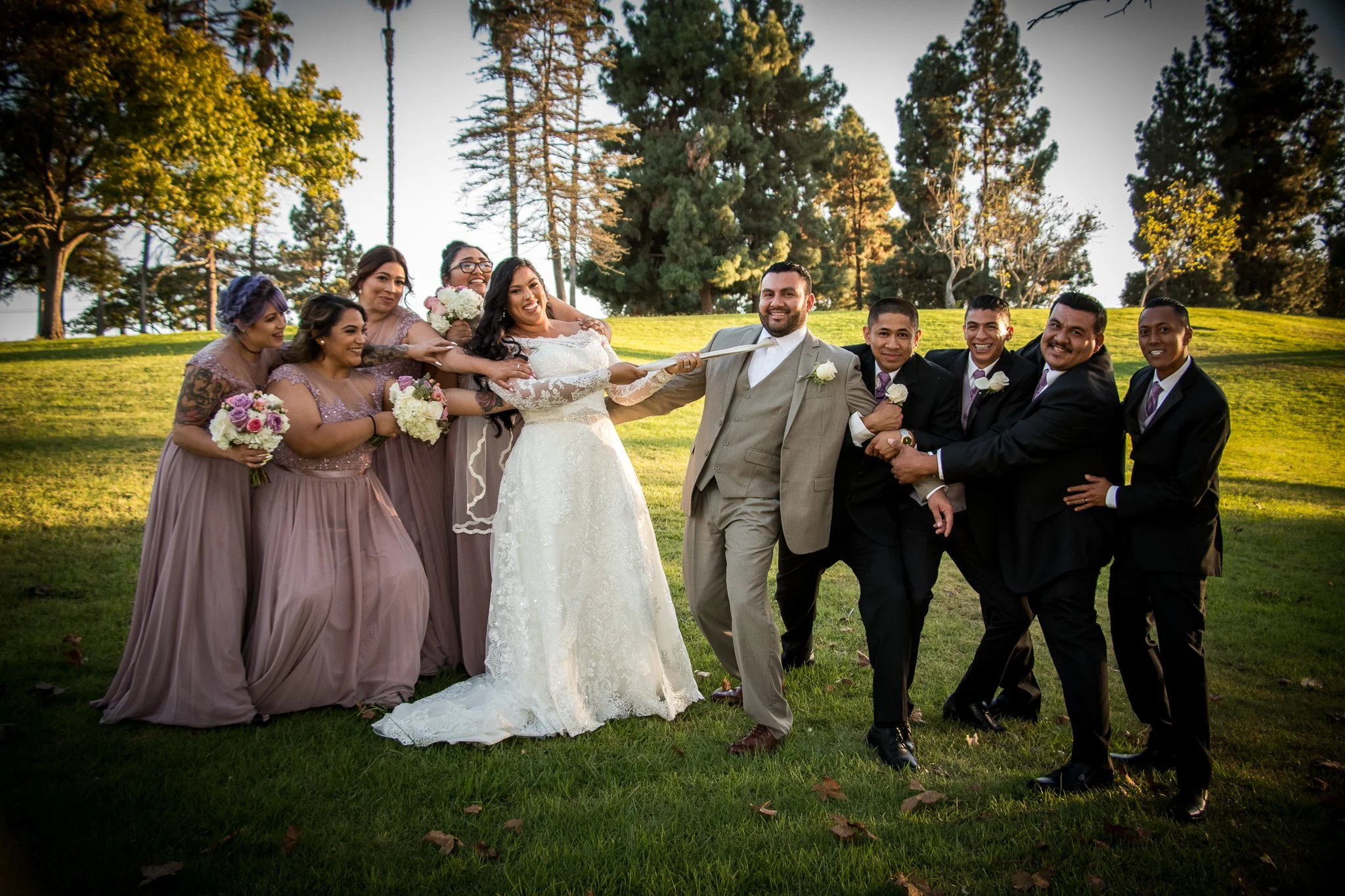 Group of people, including bride and groom, engaged in a playful tug-of-war with a ribbon outdoors on a grassy area with trees in the background.