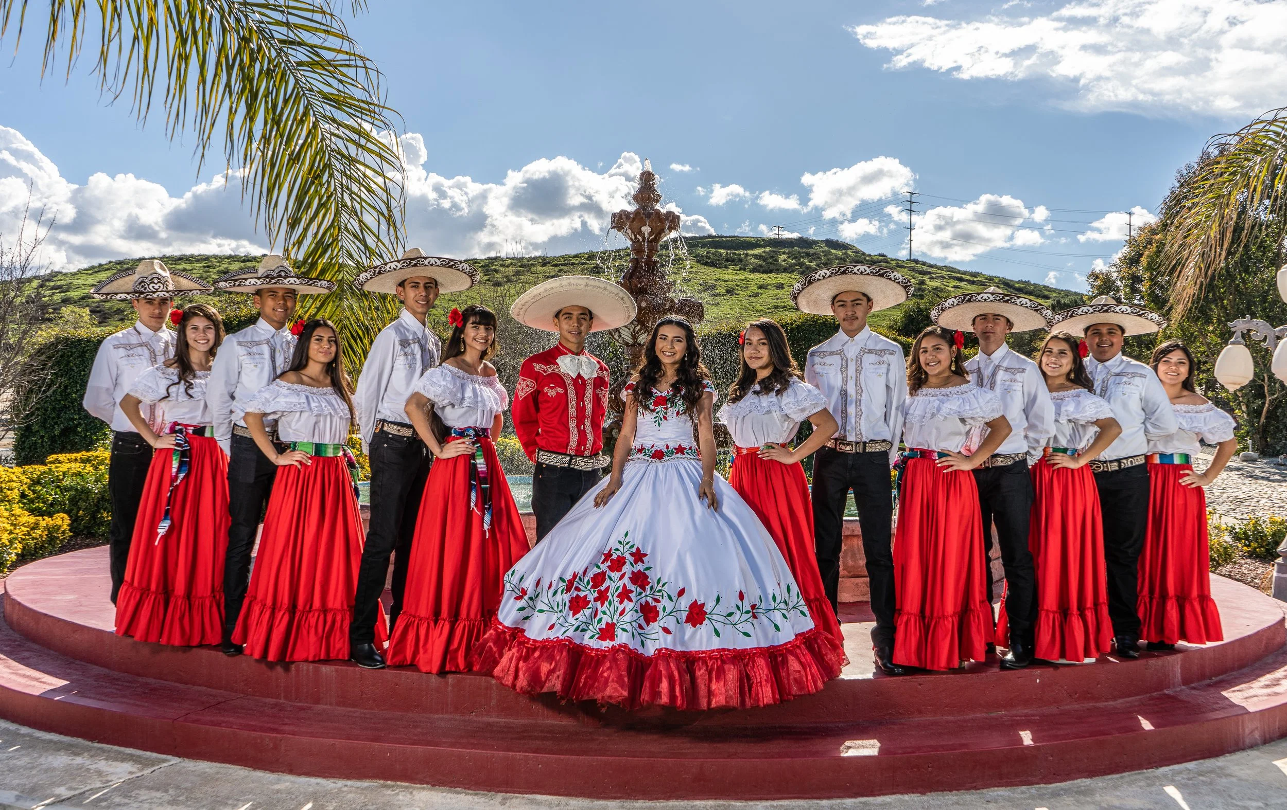 Group of people in traditional Mexican attire standing outdoors in front of a fountain, with a scenic hillside background and palm trees, during daytime.