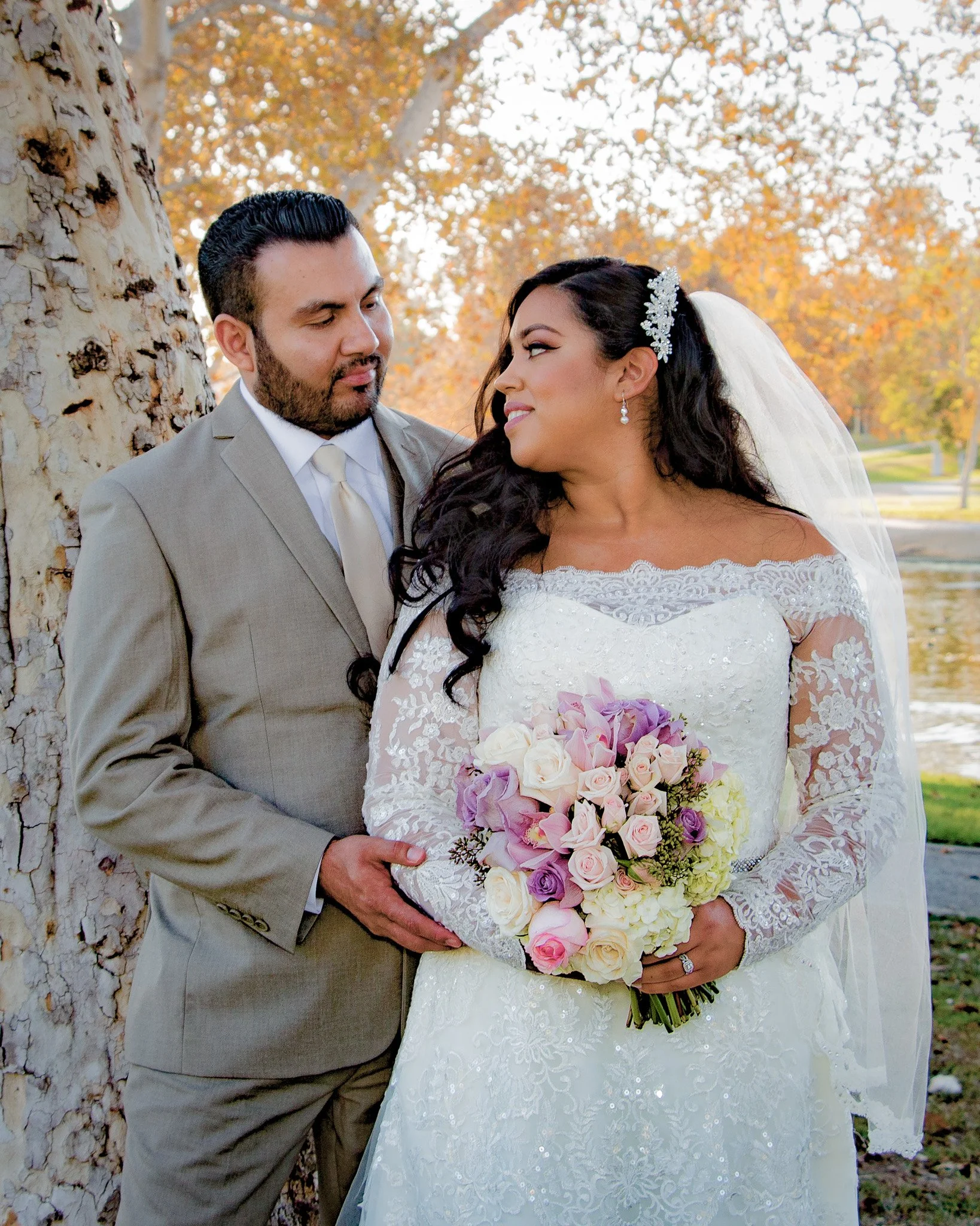 A bride and groom standing outdoors, gazing at each other, with a tree and autumn foliage in the background. The bride is holding a bouquet of pink, purple, and white flowers, wears a white wedding dress with lace sleeves, and has a veil and floral h
