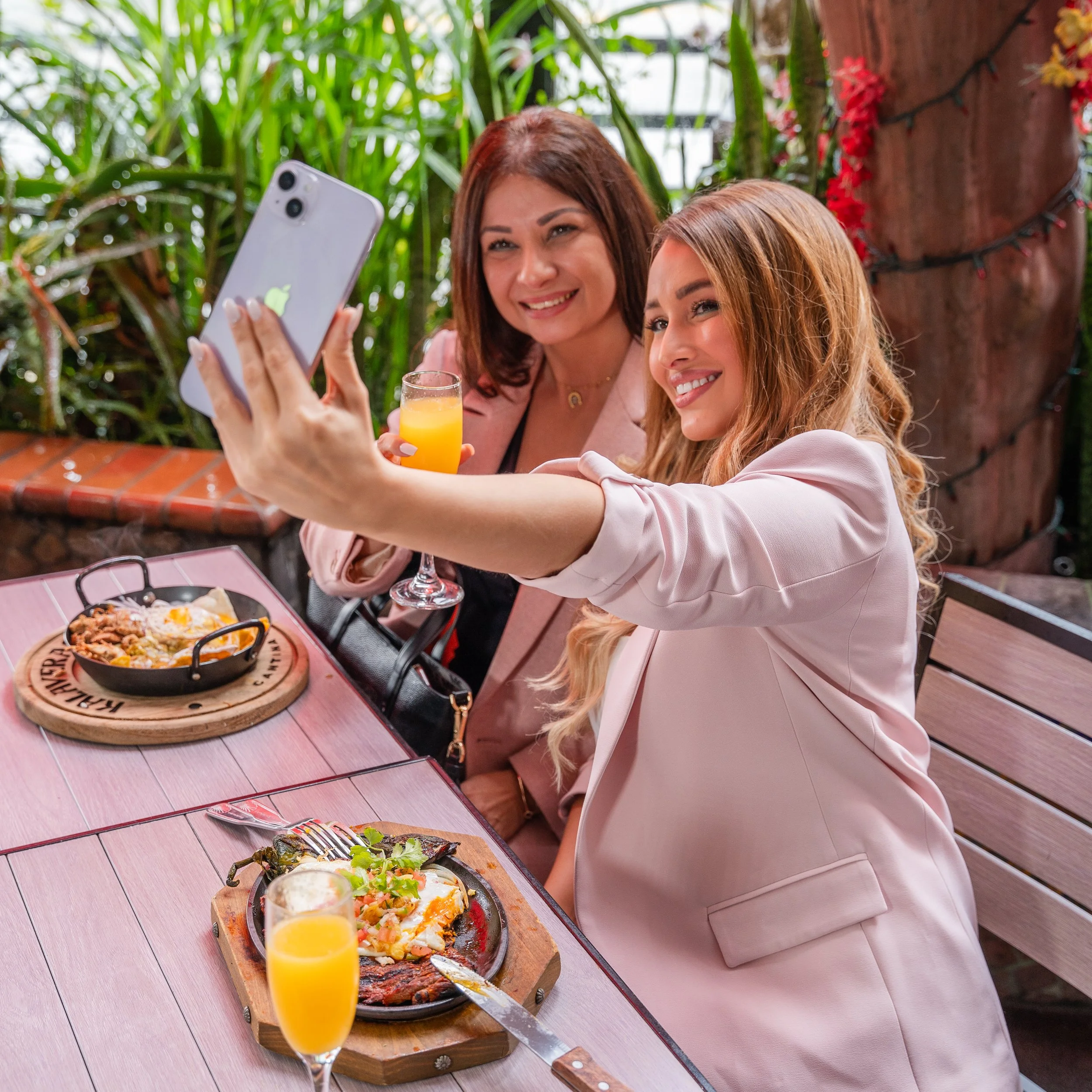 Two women taking a selfie at a restaurant table with food and drinks, surrounded by greenery and string lights.