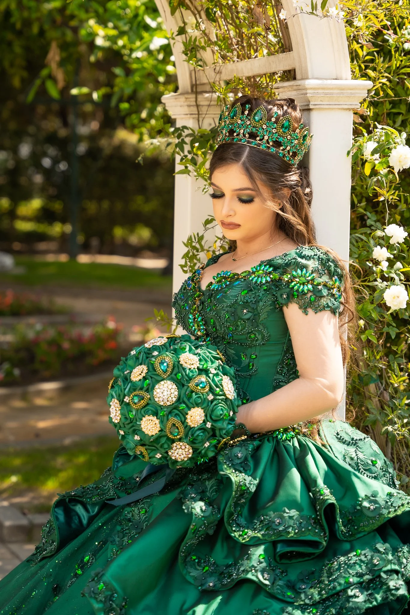 A woman dressed as a princess in a green gown with intricate embroidery and beading, wearing a large green jeweled crown, holding a bouquet of green ribbons and embellishments, standing outdoors near white flowers and greenery. Quinceañera, XV.