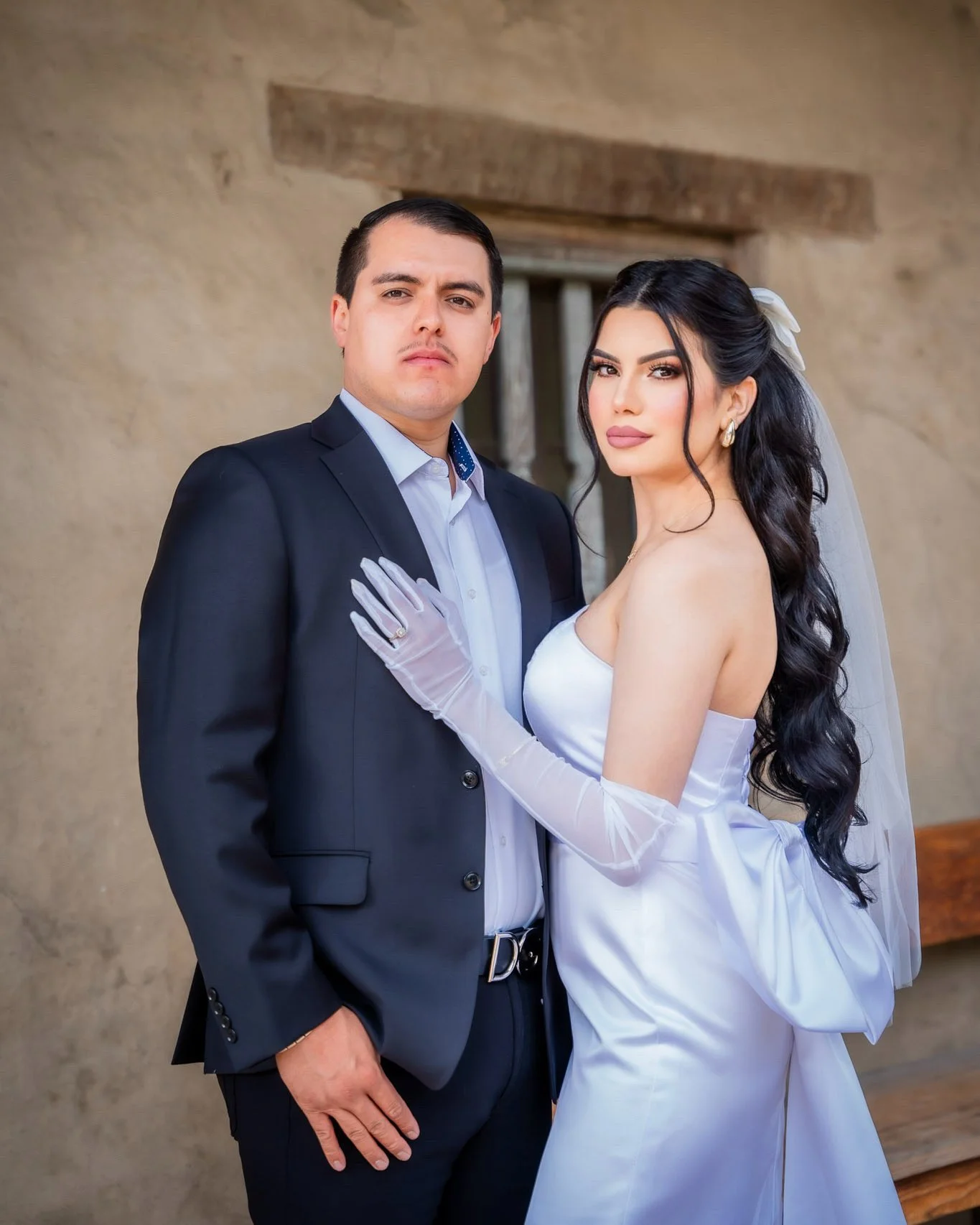 A bride and groom posing together for a wedding photo outdoors against a rustic wall. The woman has long dark wavy hair, wears a white strapless wedding gown, long white gloves, and a veil, while the man has short dark hair and is dressed in a black 