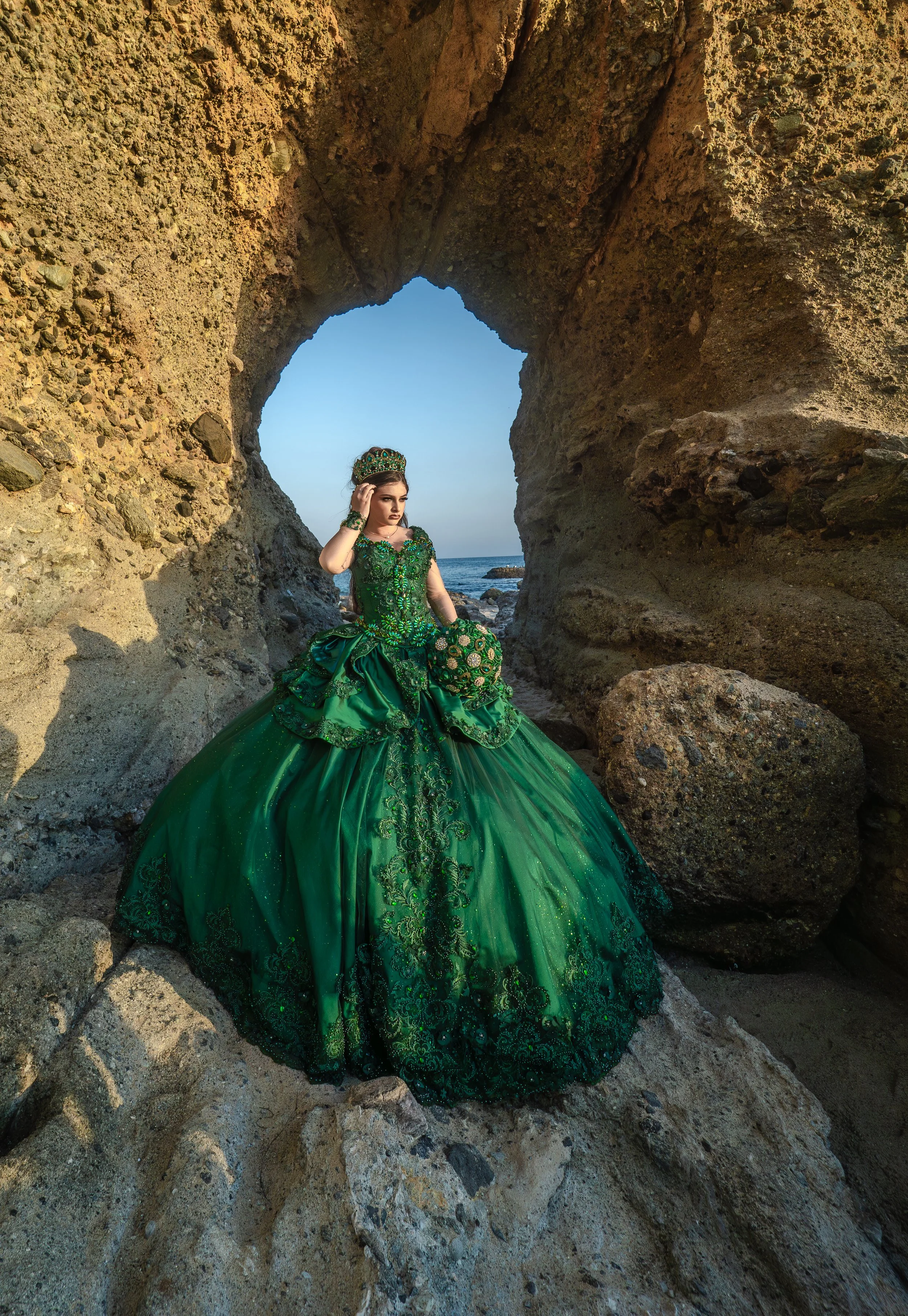 A woman wearing an elaborate green ball gown with intricate embroidery and embellishments, standing among large rocks under a natural stone arch with the ocean in the background.
