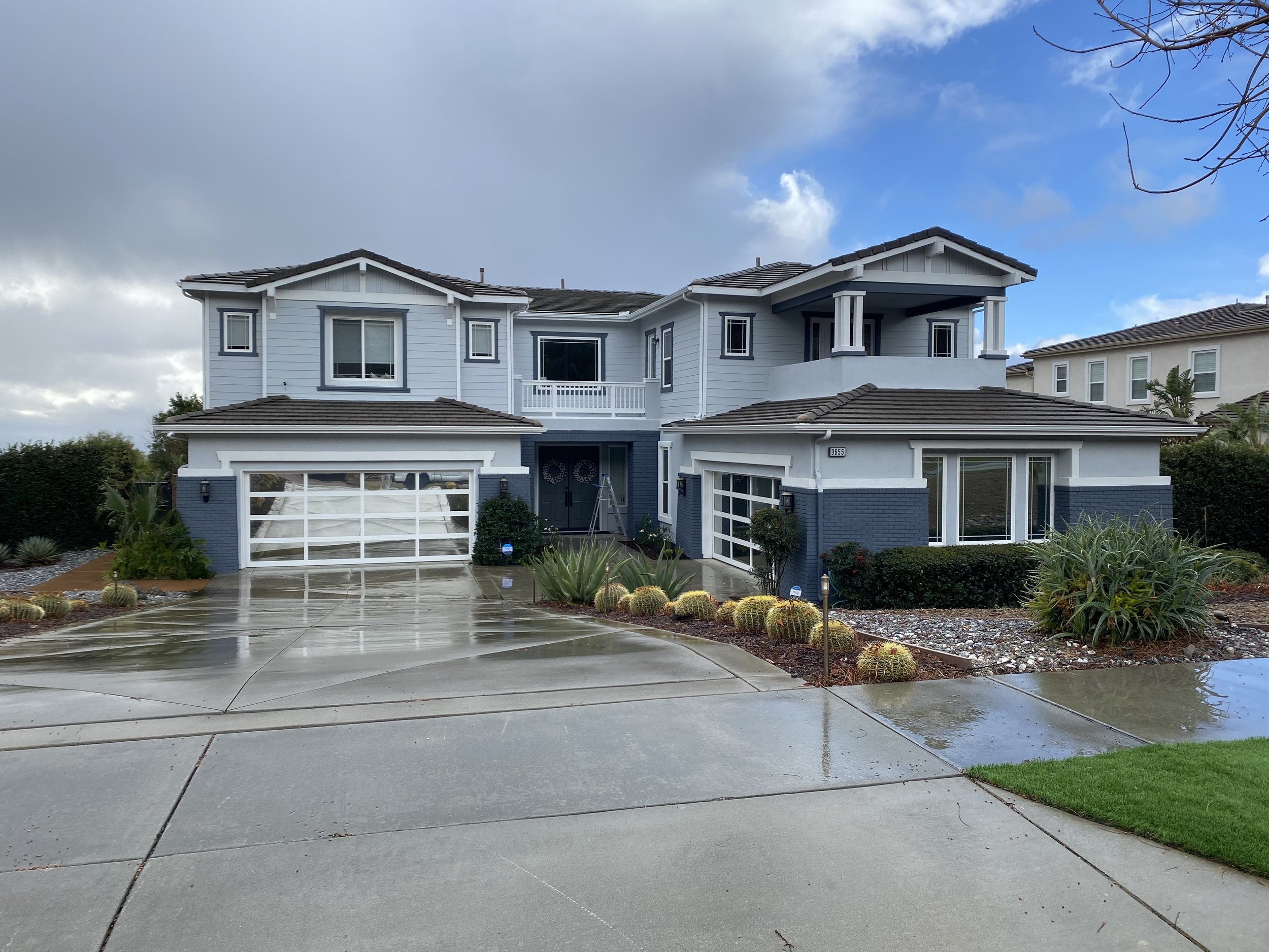 Modern two-story house with gray and white exterior, two garages with glass doors, and wet driveway showing recent rain. Landscaping includes cacti, aloe, bushes, and mulch. Overcast sky with clouds.