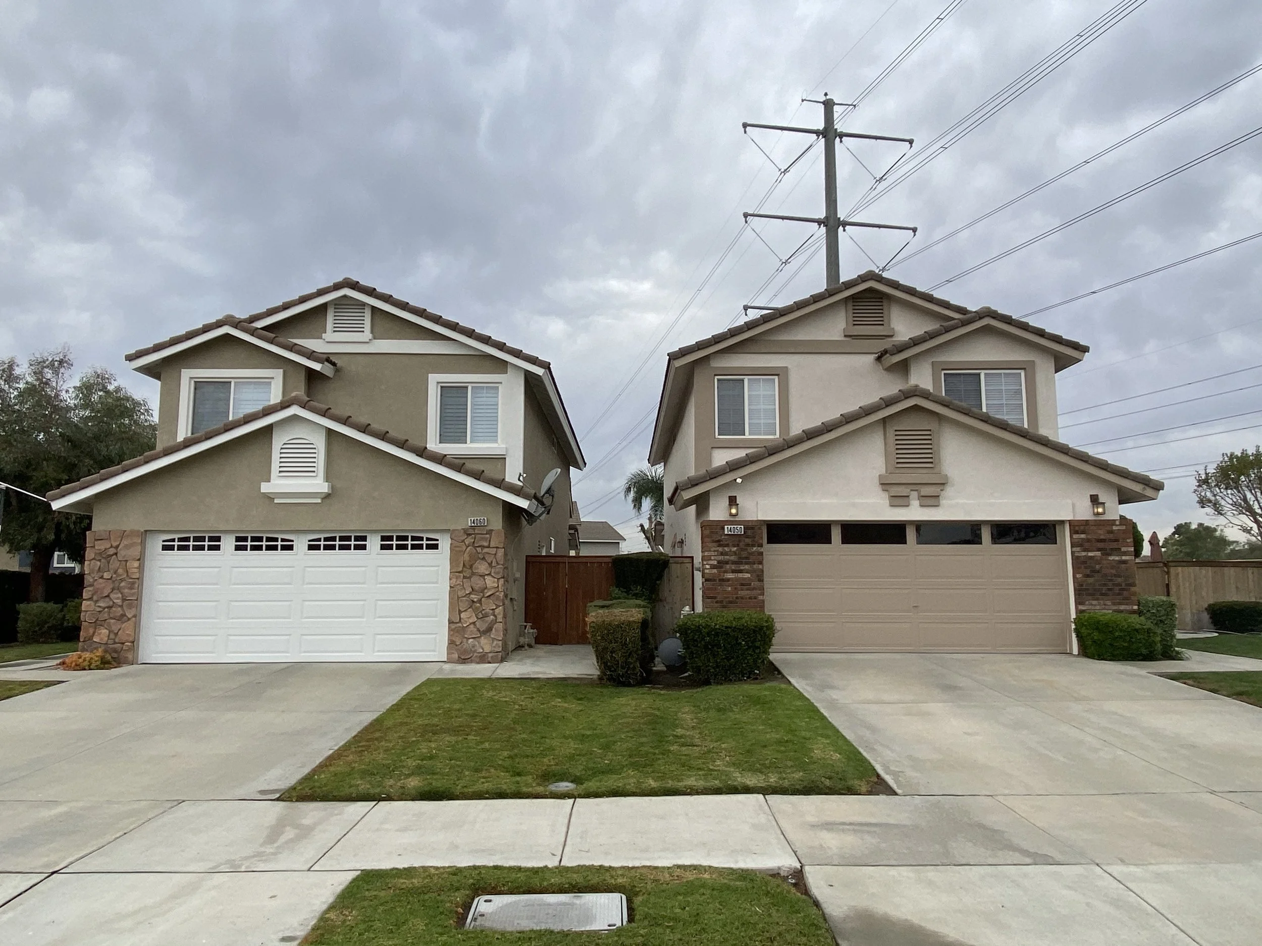 Two modern two-story houses with attached garages, front yards with grass and shrubbery, overcast sky, power lines overhead.