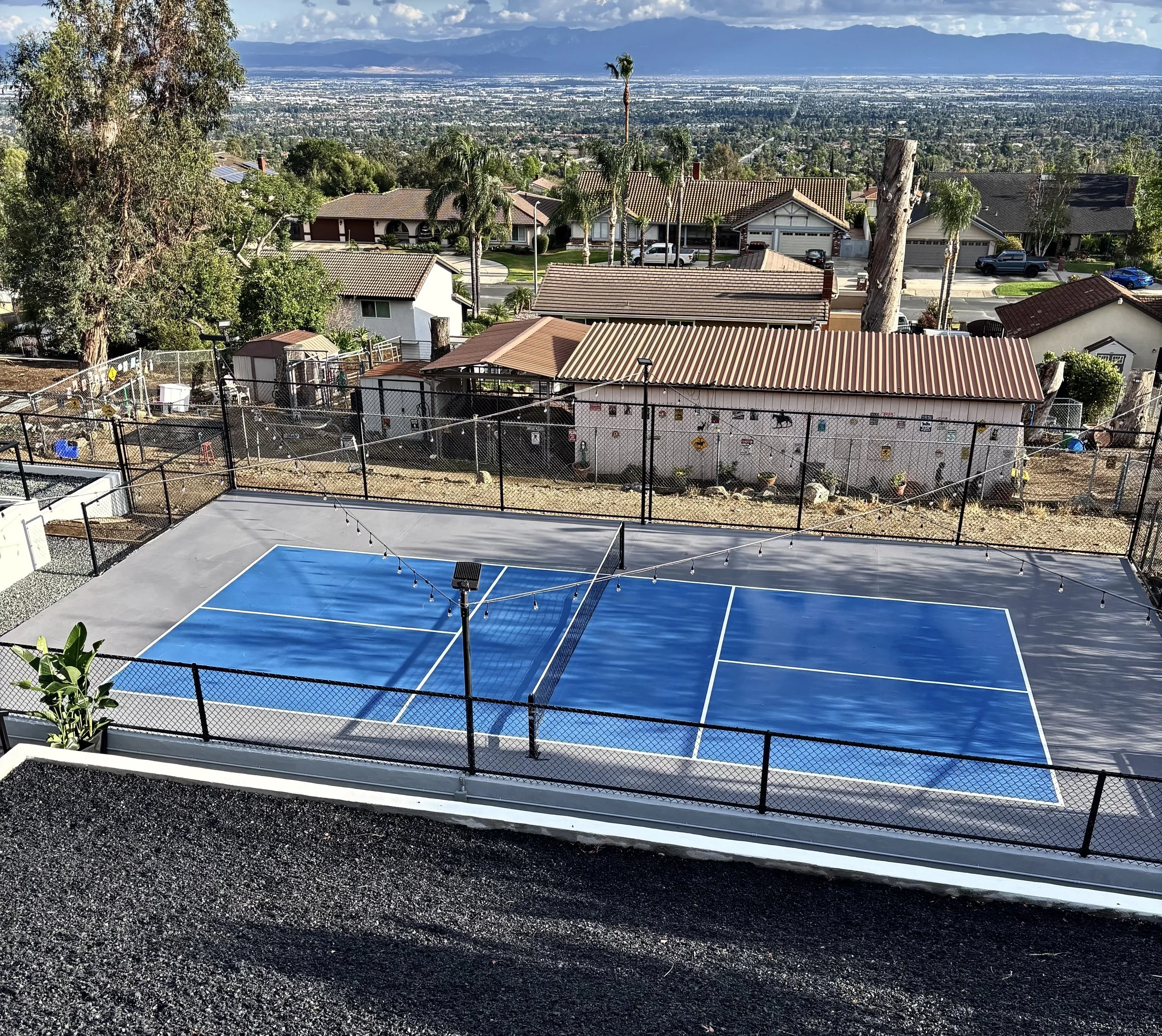 Third-floor view of a blue tennis court with string lights and a solar-powered light, surrounded by a black chain-link fence; suburban neighborhood with houses, trees, and distant mountains in the background.