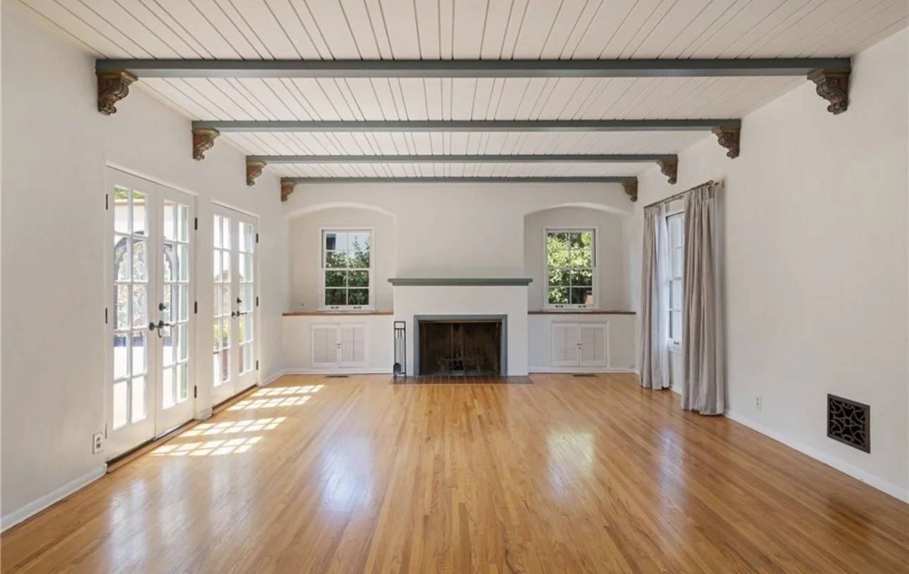 Empty living room with hardwood floors, white walls, a fireplace, three windows, French doors, and curtains.