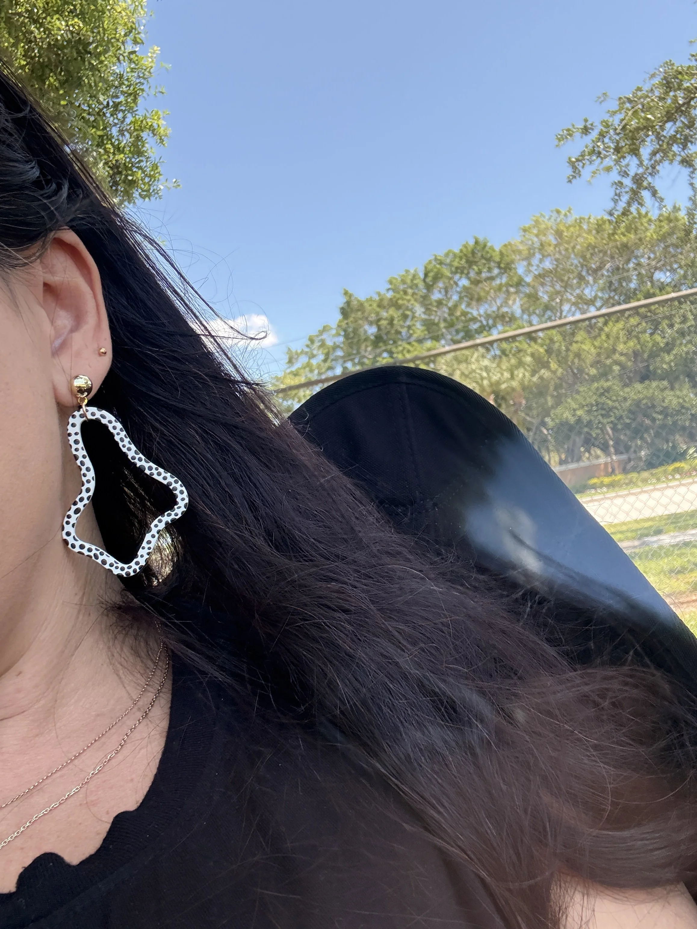 A close-up of a woman wearing dangling earrings, a black top, and layered gold necklaces, with long dark hair, outdoors under a blue sky with trees and a fence in the background.