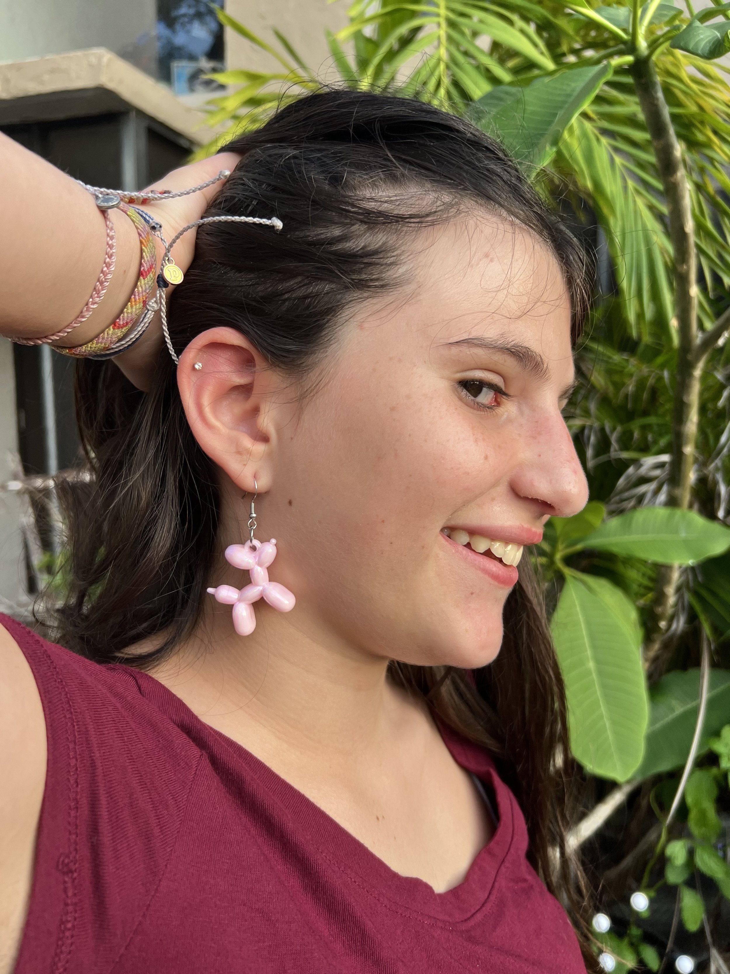 A woman smiling and showing her profile, wearing a large pink balloon flower earring, a multicolored bracelet, and a small silver earring, with lush green plants in the background.