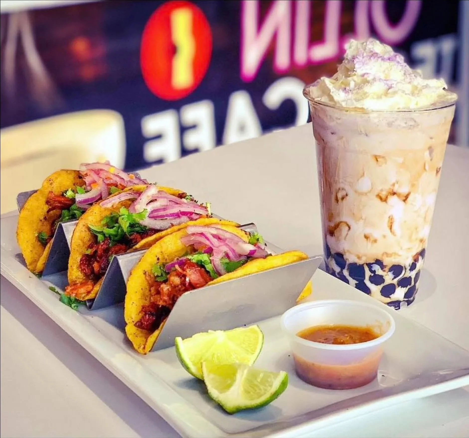 Three tacos topped with onions and cilantro on a tray with lemon wedges, a cup of salsa, a tall glass of a pink and white blended beverage topped with whipped cream, and a bubble tea with tapioca pearls.