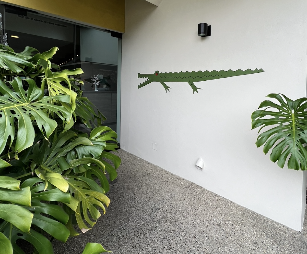 Green cartoon crocodile wall decoration on white wall near large green monstera plants.