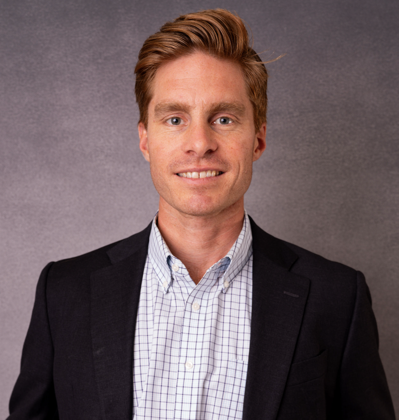 A young man with red hair, blue eyes, wearing a dark blazer and a light checkered shirt, smiling at the camera against a gray background.