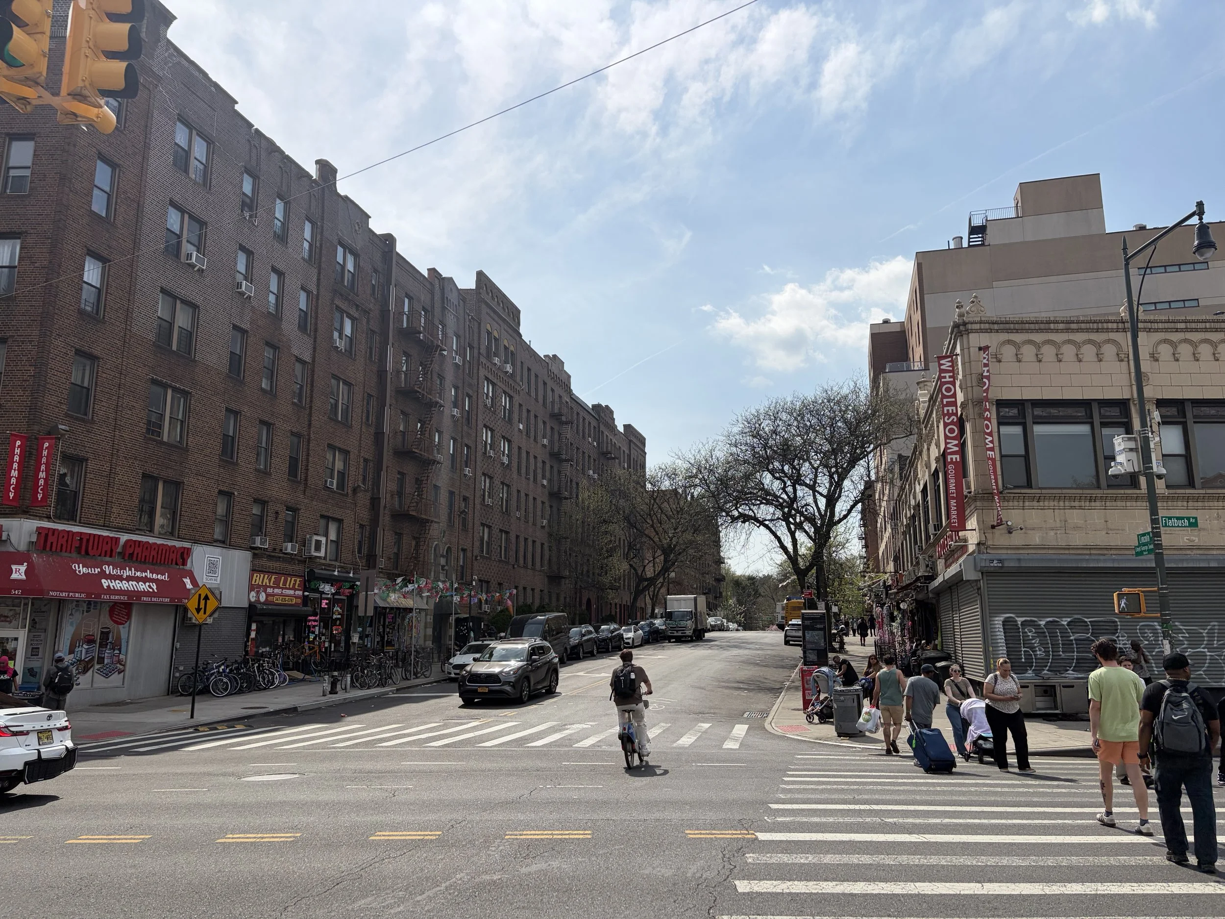 A busy city street with people walking, cycling, and cars parked along the curb. Tall brick buildings with storefronts and signage, and a streetlight visible. The sky is partly cloudy.