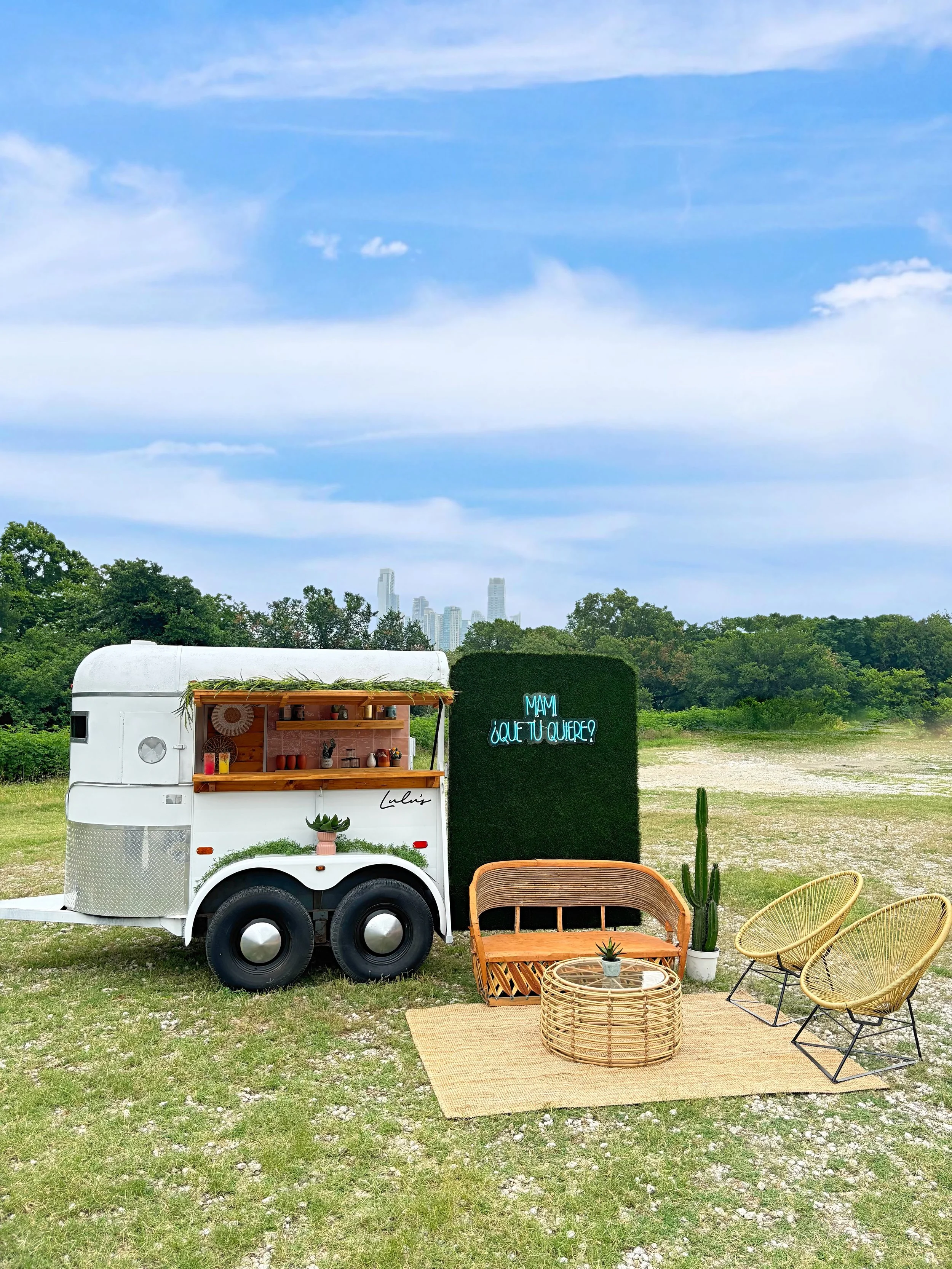 A white food truck with a wooden shelf along the side, in an outdoor setting with chairs, a rug, and a cactus, with trees and a city skyline in the background and a blue sky with clouds.