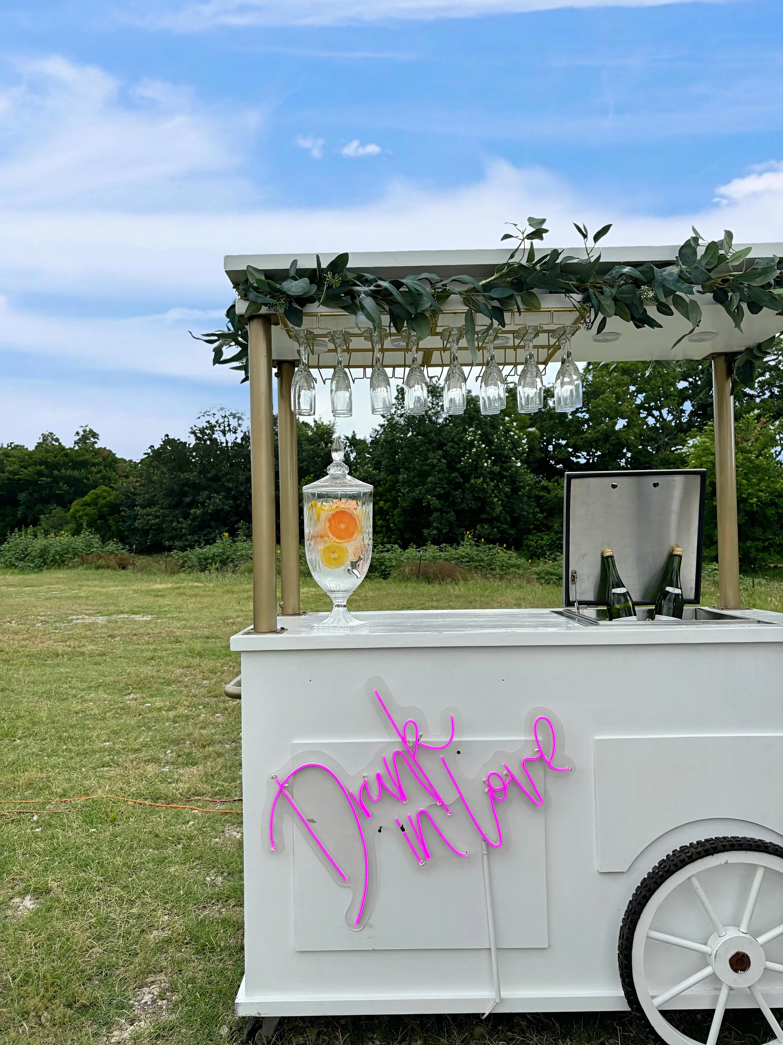 A white mobile bar cart decorated with greenery and hanging wine glasses, set outdoors on grass with trees and a blue sky in the background, featuring a neon pink sign that says 'Drink'.