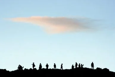 Silhouetted hikers and people standing on a hilltop against a blue sky with a few clouds.