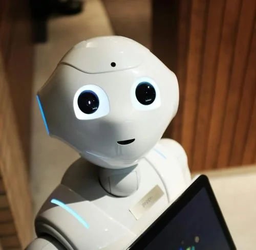A friendly robot with a white, rounded head and large, expressive eyes looking at a tablet.