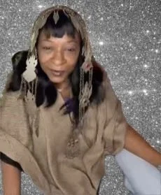 Woman with long dark hair and braids, wearing a brown and beige outfit, standing in front of a sparkly silver background.