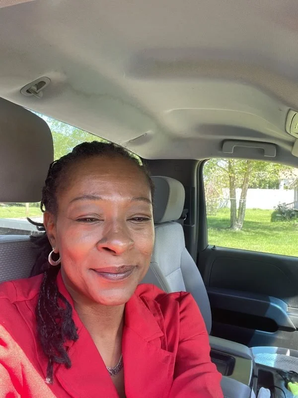 A woman with braided hair and hoop earrings taking a selfie inside a vehicle, with a window showing green trees and grass outside.