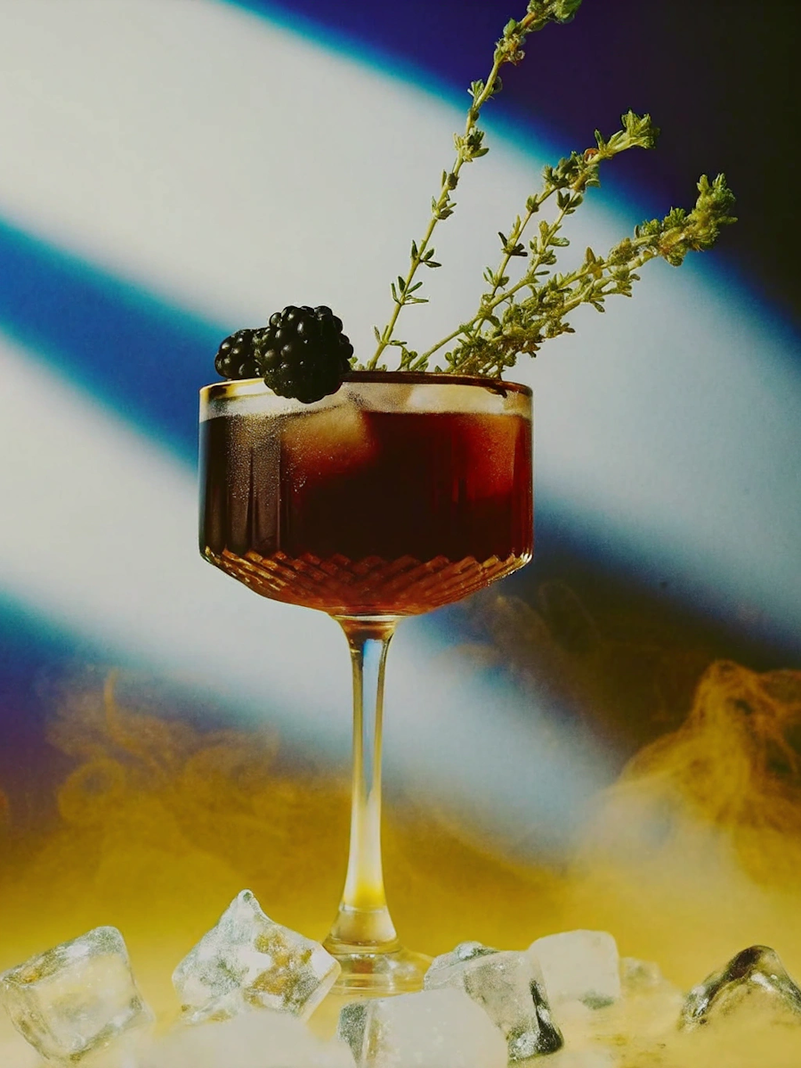 A cocktail glass with dark red drink garnished with blackberries and rosemary sprigs, surrounded by ice cubes on a surface with a blurred mountain background.