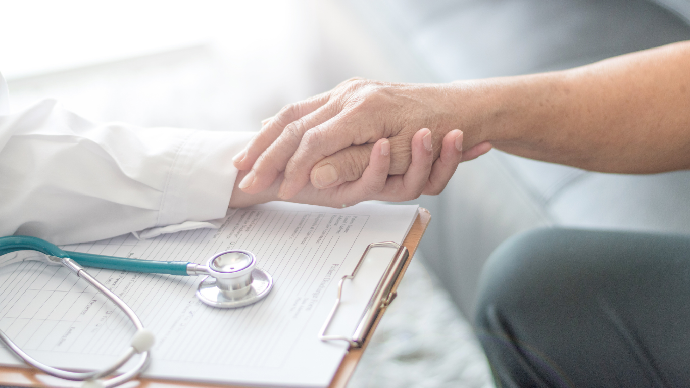 A healthcare professional holding a patient's hand, with a stethoscope and medical chart on a clipboard nearby.