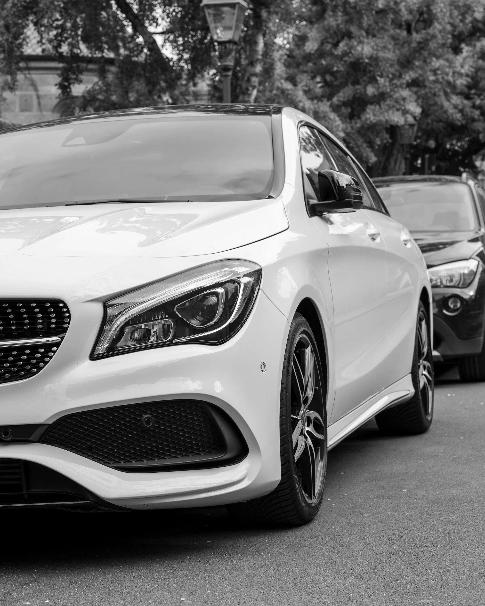 Close-up of the front part of a white modern sports car parked outdoors, with another vehicle and trees in the background.