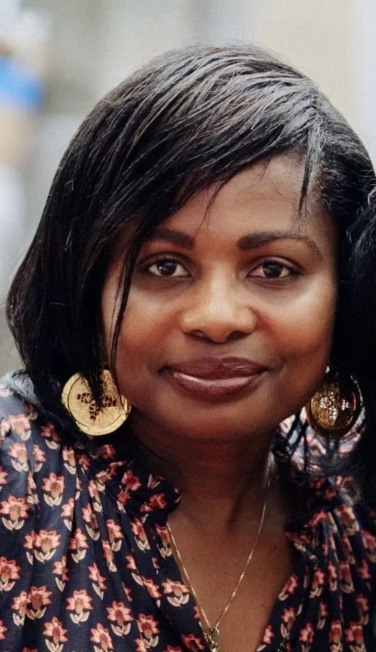 A woman with shoulder-length black hair, wearing large gold earrings, a floral blouse, and a necklace, smiling at the camera.