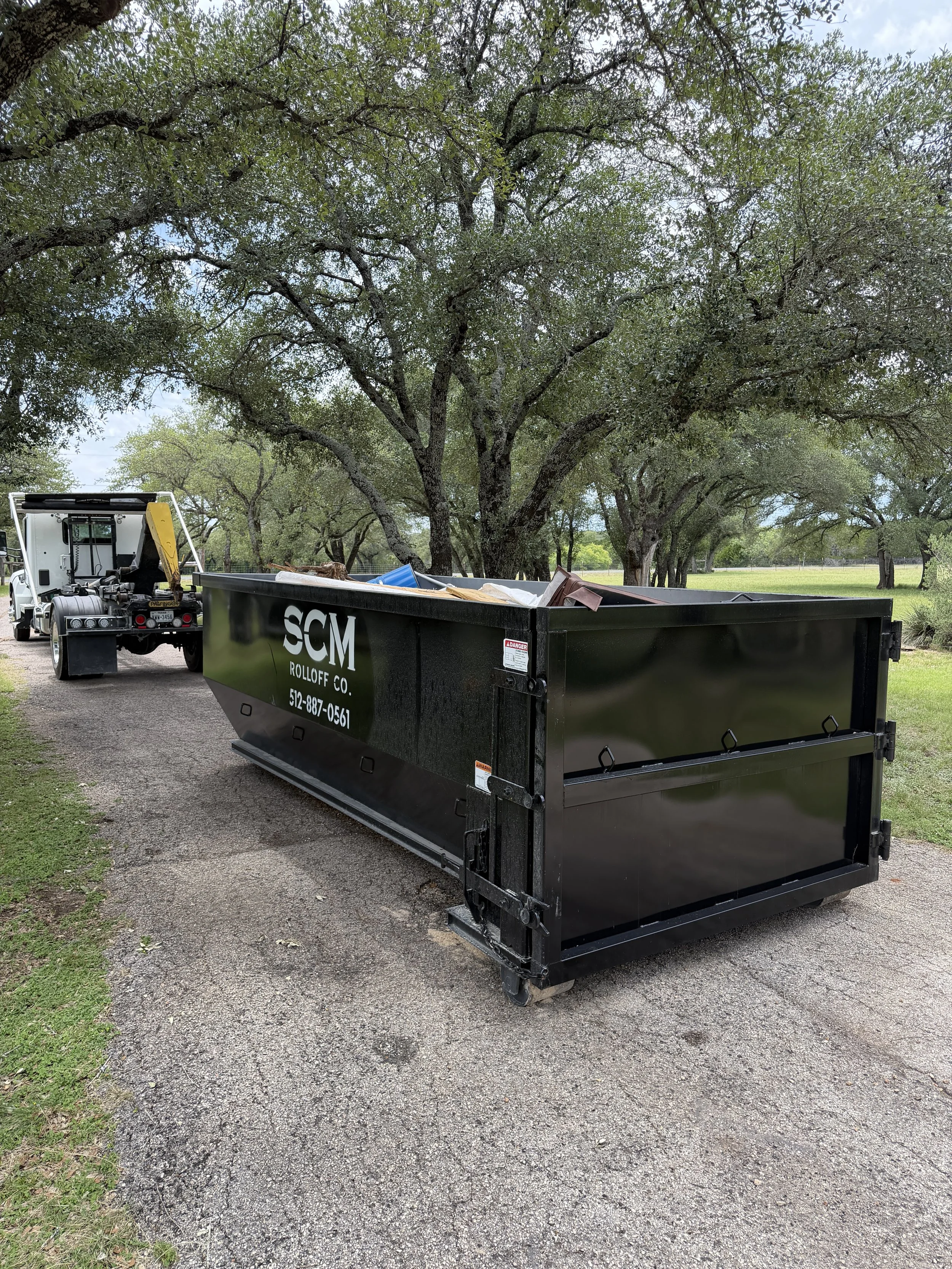 A flatbed truck filled with trash and debris parked on a gravel path beneath a canopy of large trees in a park or rural area.