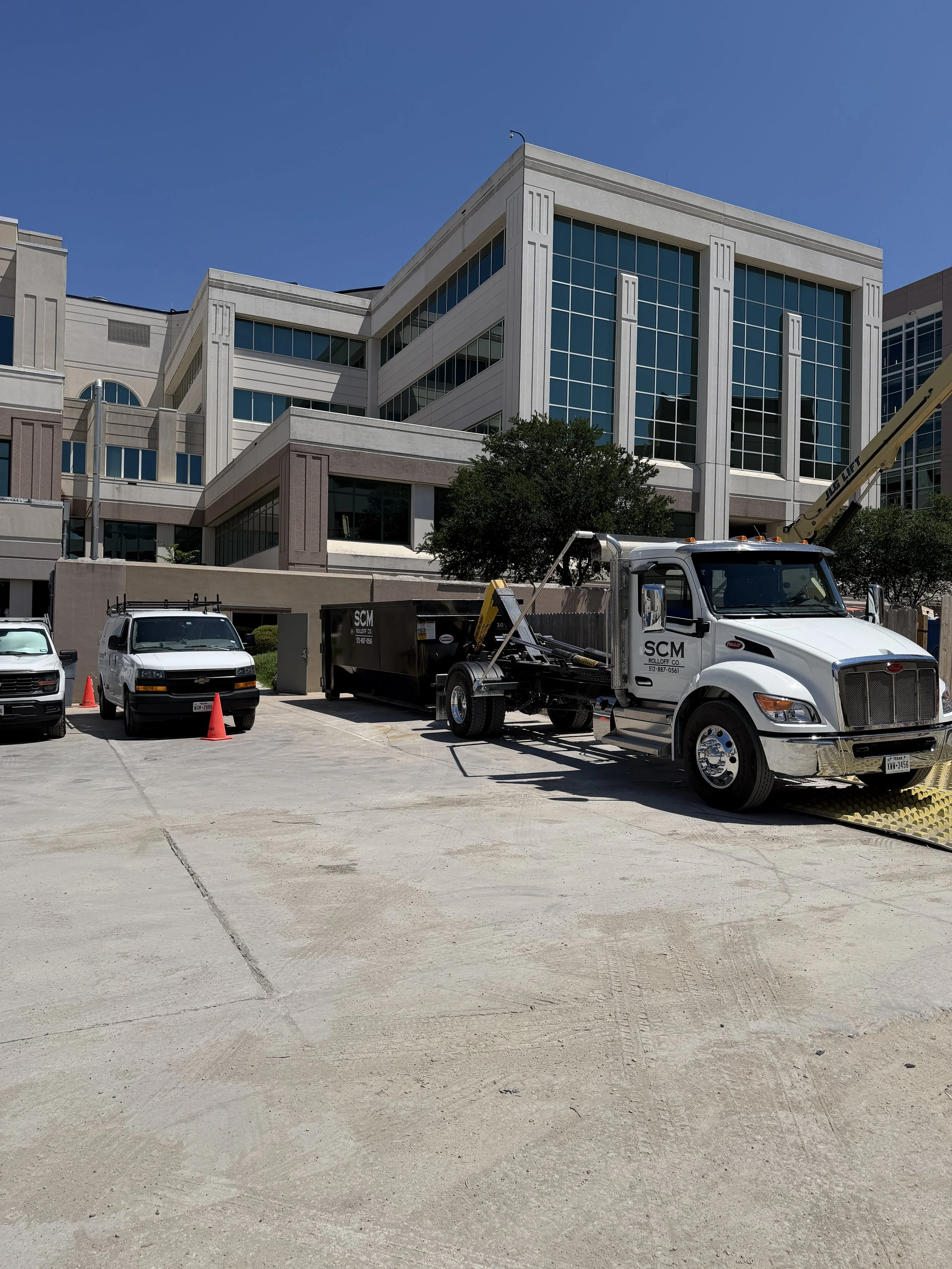 A multi-story office building with large glass windows and a concrete facade. There is a white truck with an extended crane in the foreground, along with several parked white vehicles and orange cones in a parking lot.