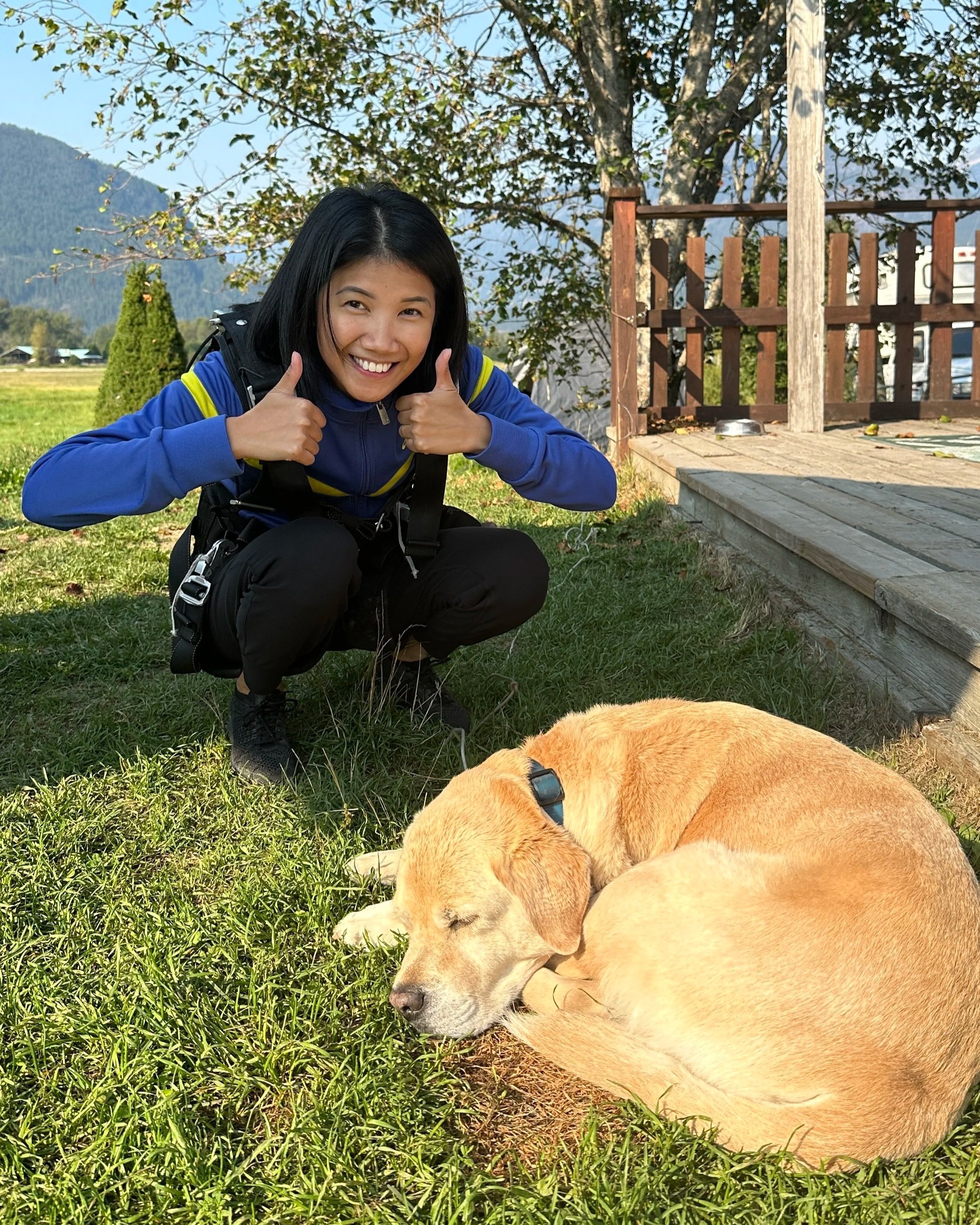 A woman crouching on the grass giving two thumbs up, smiling, next to a sleeping yellow Labrador Retriever dog outdoors with a wooden deck, trees, and mountains in the background.