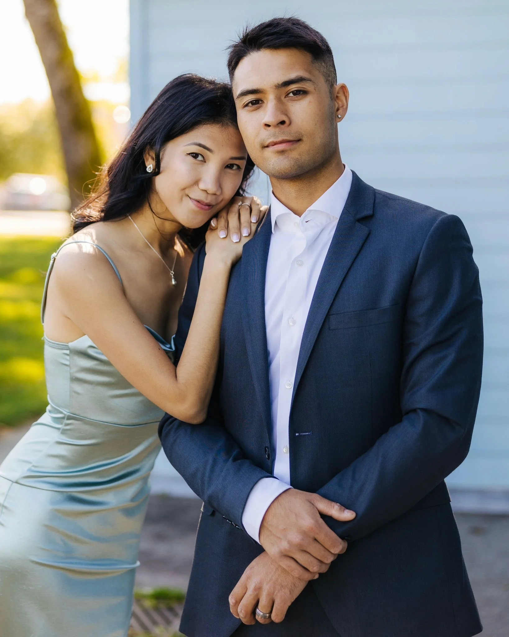 A young woman with dark hair and a satin dress stands beside a young man in a suit, both posing outdoors with a house and trees in the background.