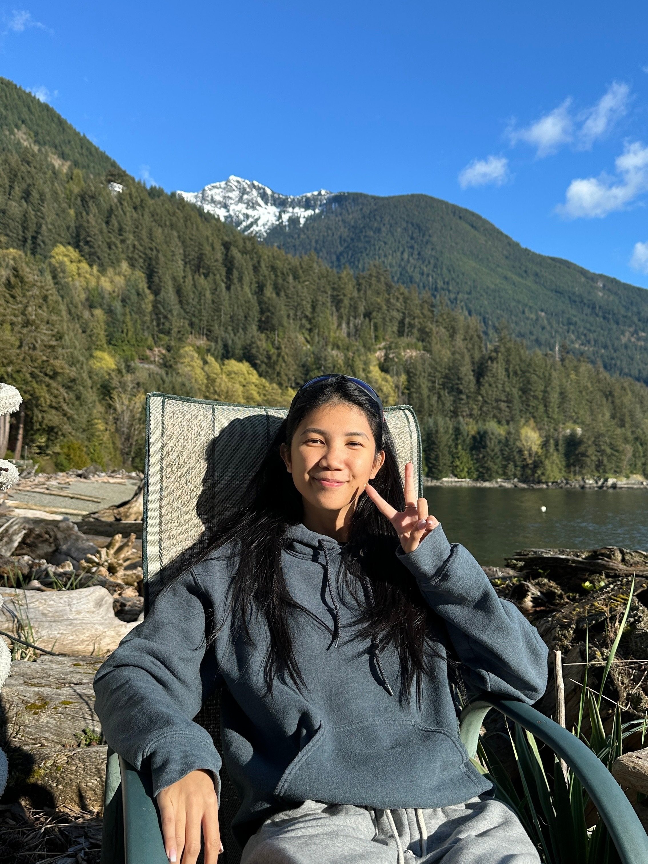 A young woman sitting outdoors on a reclined chair, making a peace sign with her hand, in front of a scenic mountain landscape with trees, a lake, and snow-capped peaks under a blue sky.