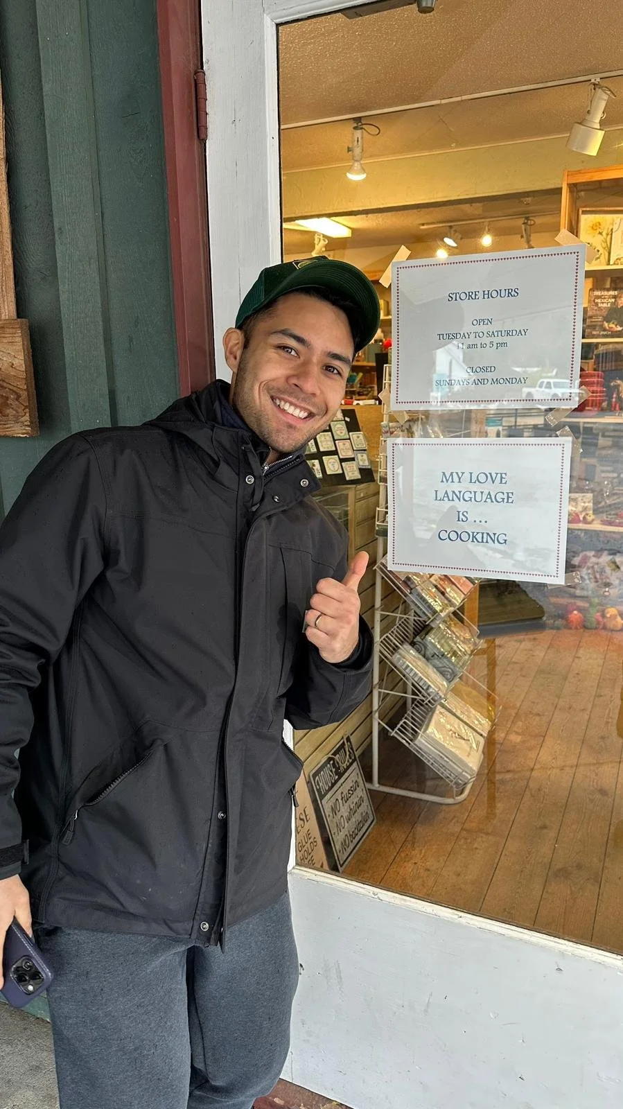 A young man with a big smile wearing a black jacket and green cap standing outside a store, pointing to a sign that says "My love language is... cooking" on the store window.