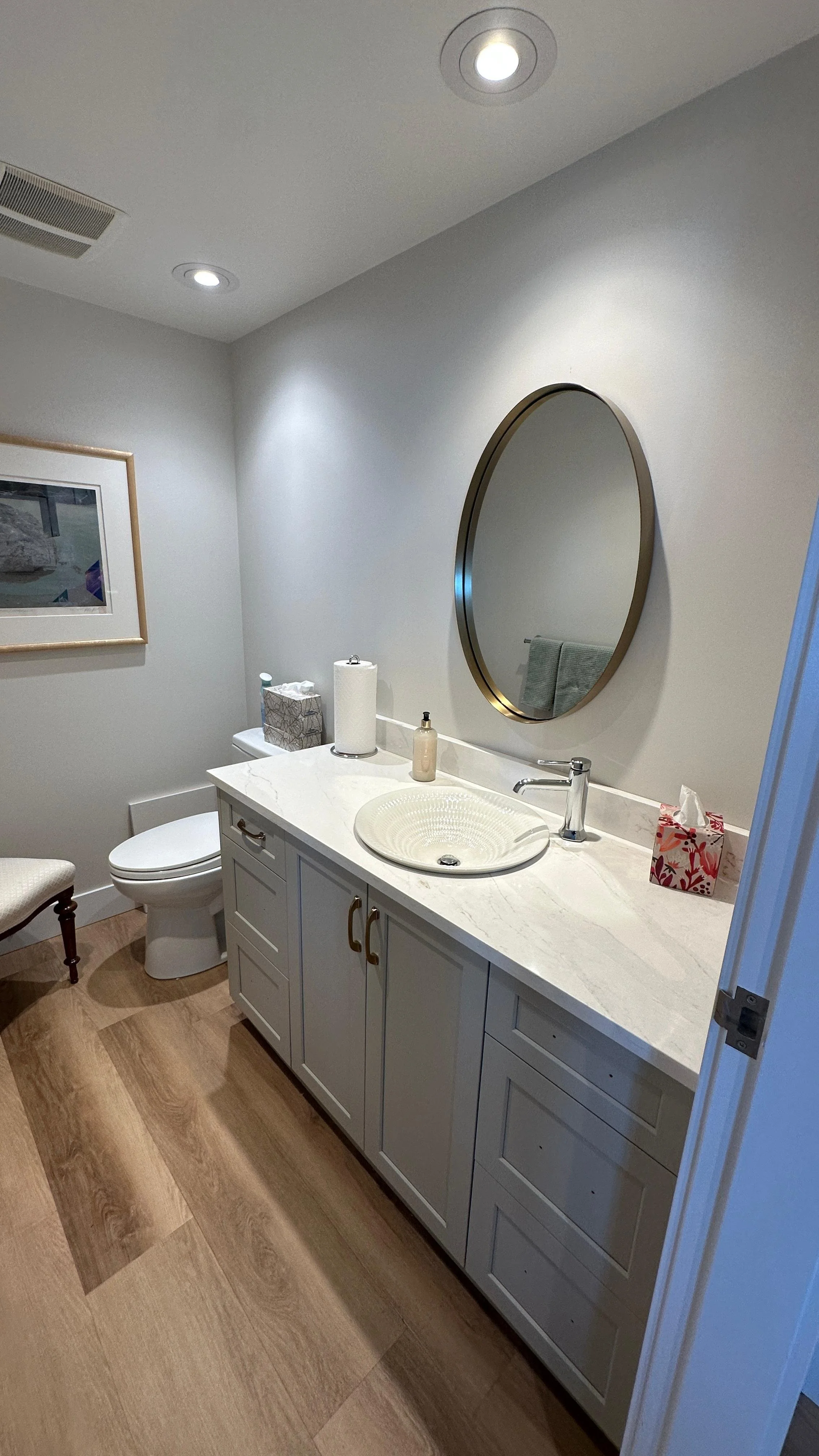 A bathroom with a white vanity, round vessel sink, mirror, tissue box, paper towels, soap dispenser, toilet, and framed picture on the wall.