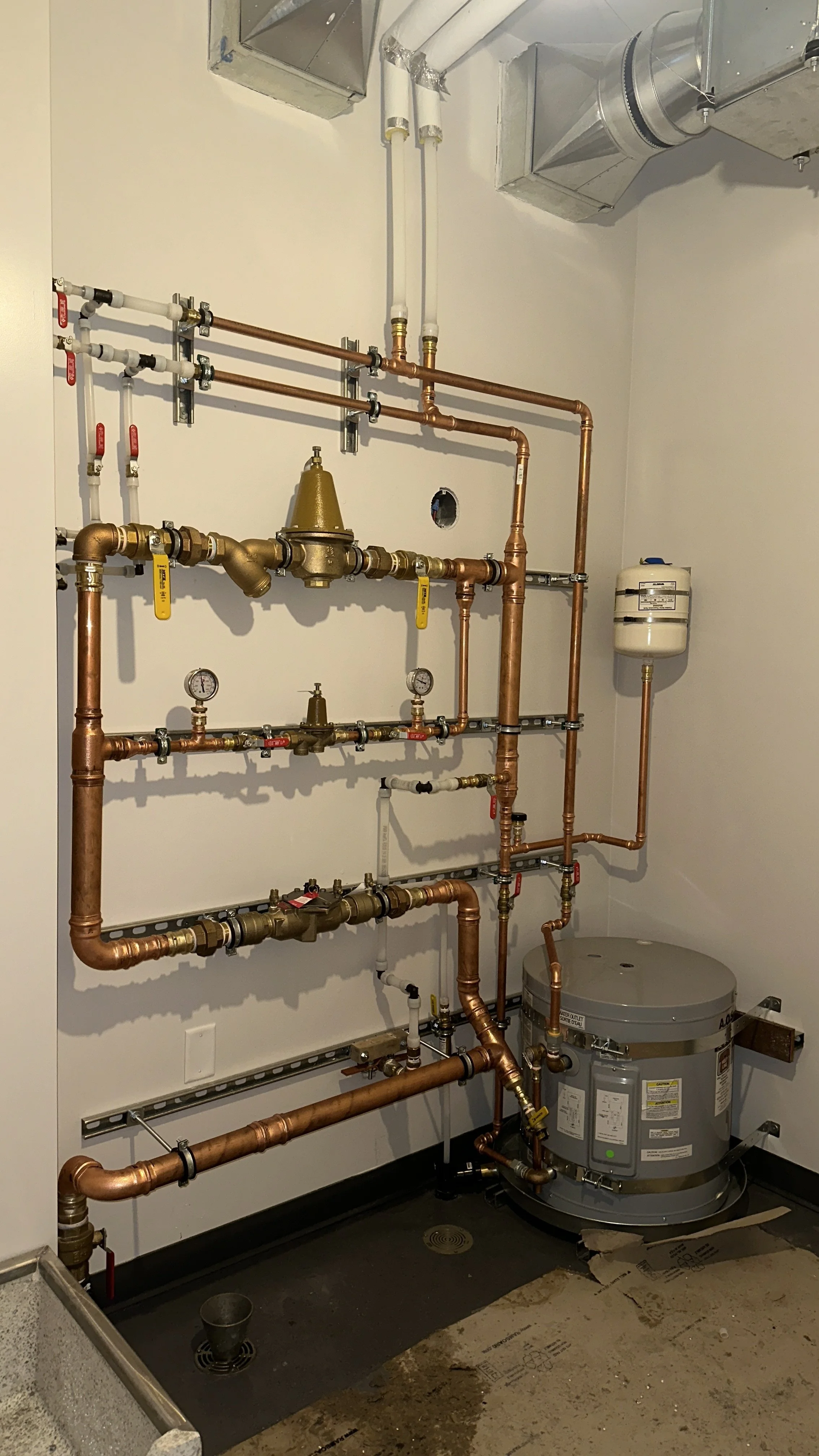 A wall of plumbing pipes and gauges in a utility room, including copper pipes, valves, and meters, with a water heater at the bottom right.