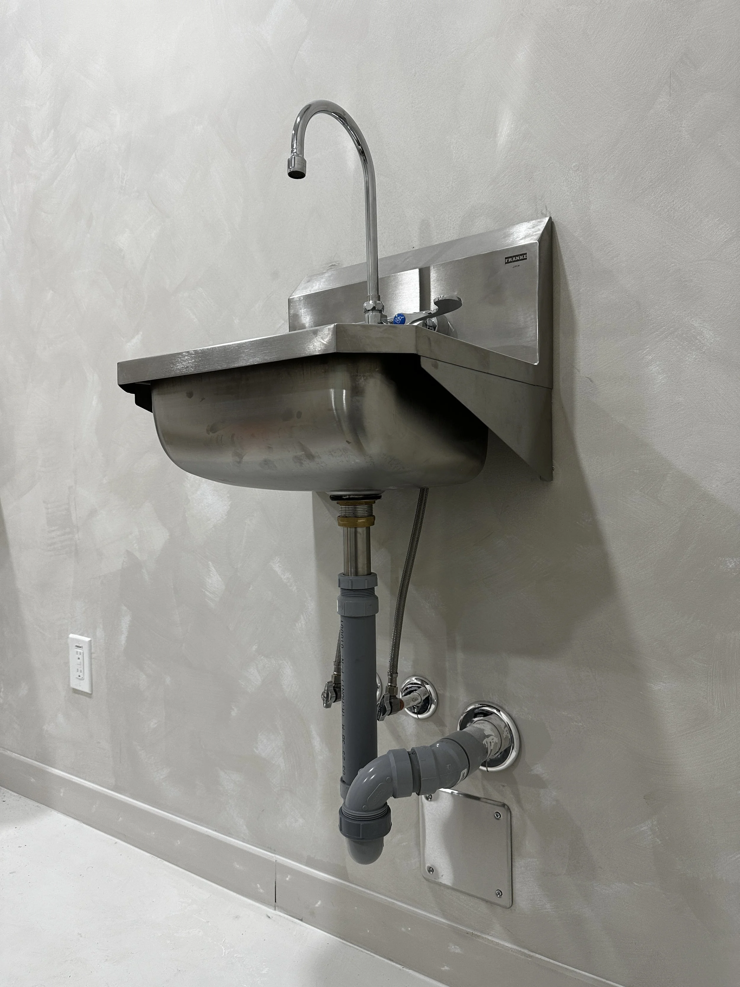 A stainless steel utility sink with a high curved faucet, mounted on a beige wall with exposed gray plumbing pipes below.