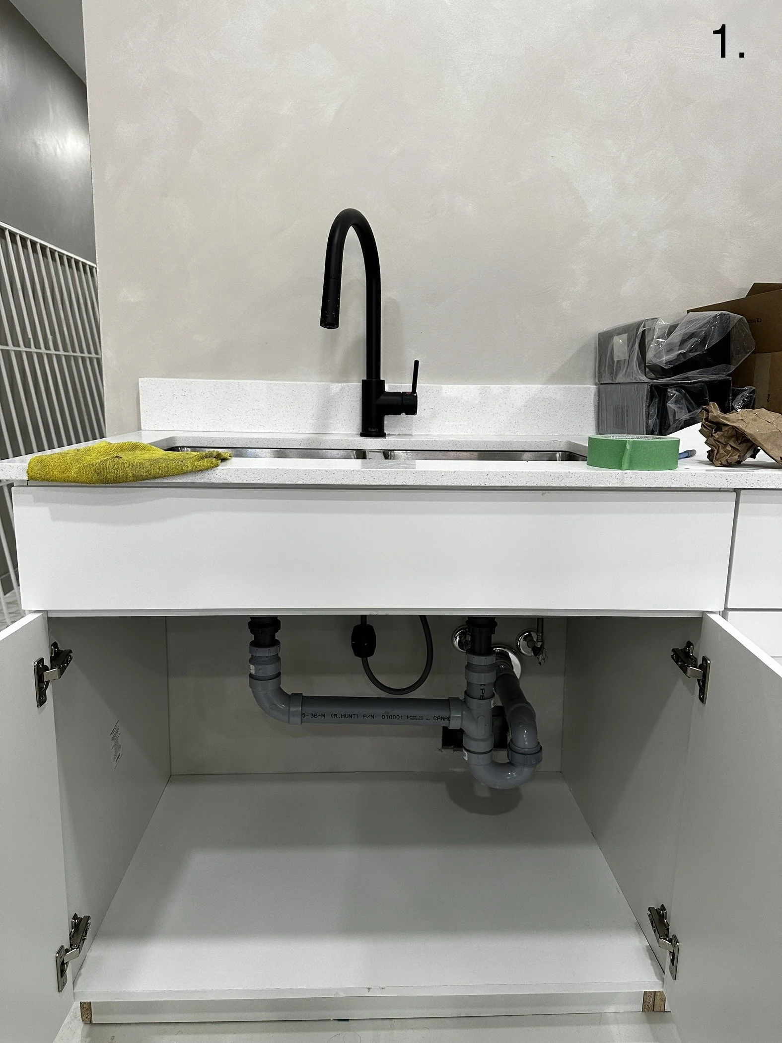 Photo of a kitchen sink with black faucet, yellow cleaning cloth on left, green painter's tape and brown paper on right, under-sink plumbing with gray pipes, open white cabinets, and a beige wall.