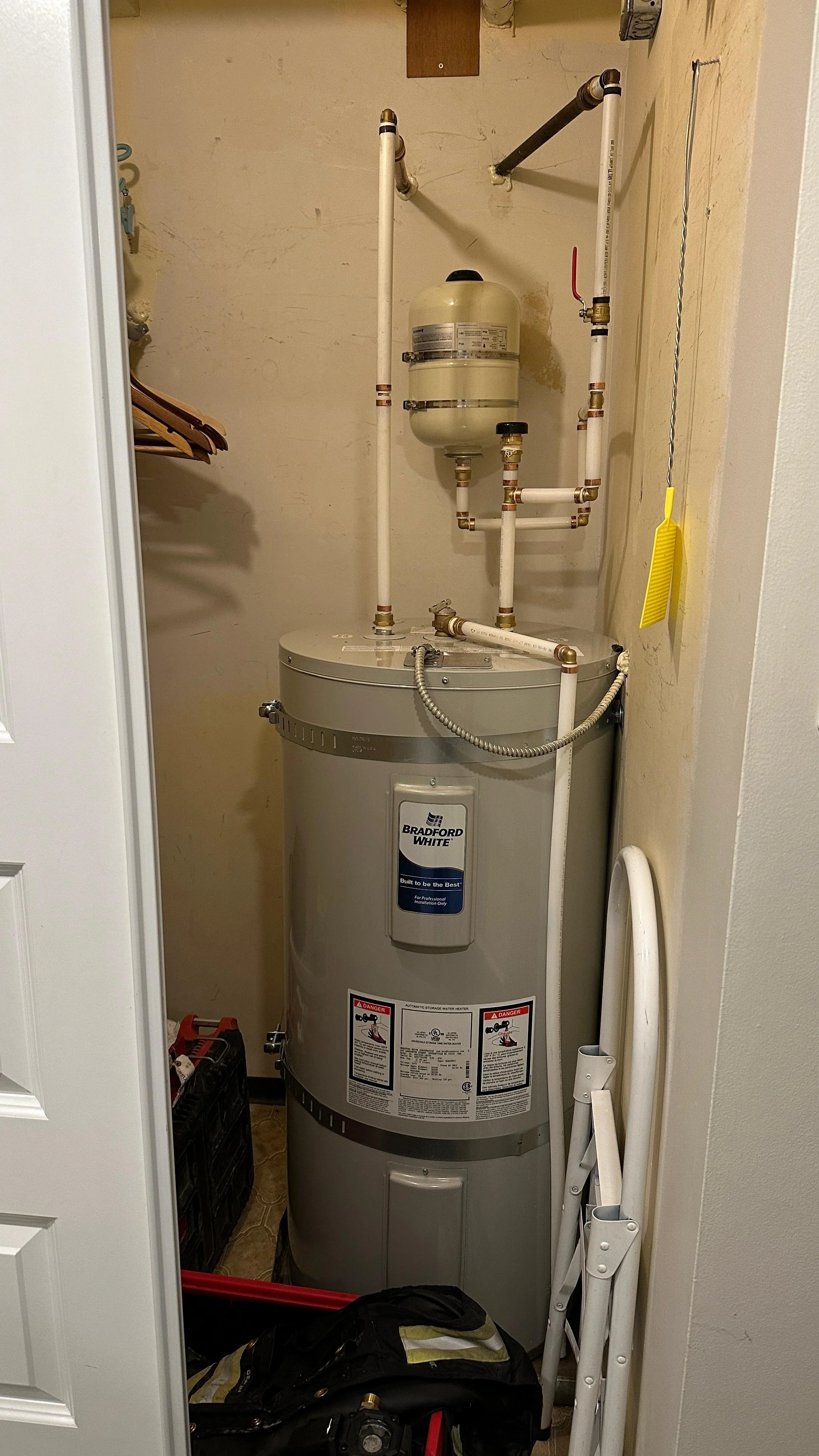 A water heater in a small closet with plumbing and a vent pipe, surrounded by tools and equipment.