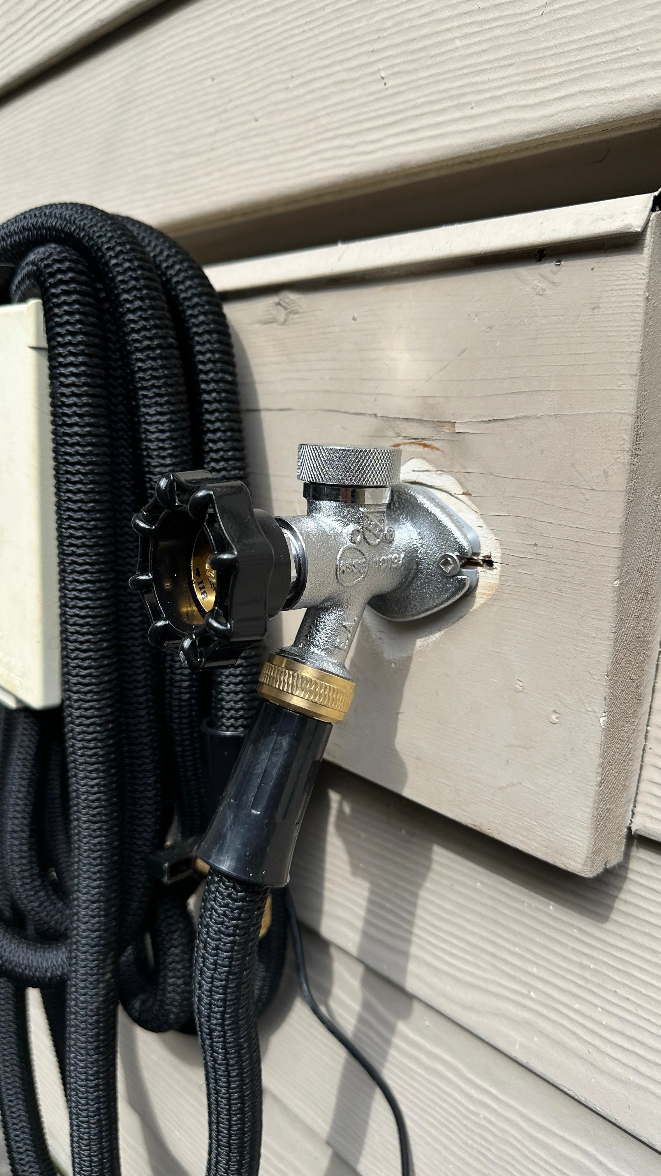 Close-up of a hose faucet connection on an exterior wall with black braided hoses and a white shed siding in the background.