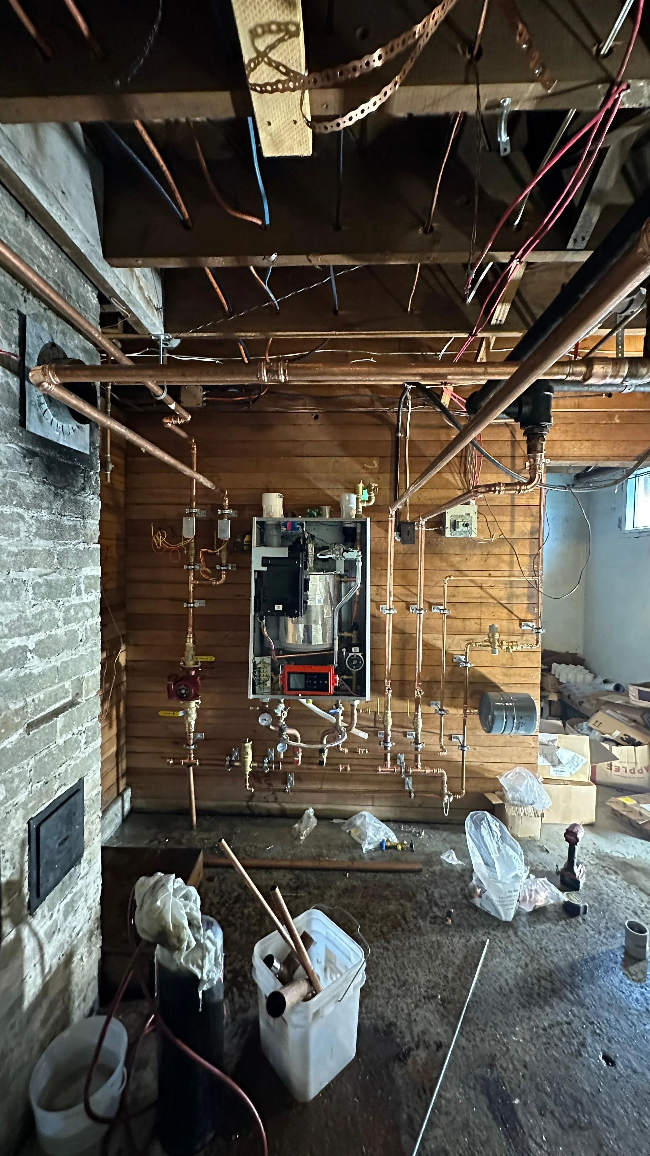 A basement with exposed copper plumbing pipes and an unfinished wall, showing a water heater or boiler installation in progress, with tools and construction materials scattered on the floor.