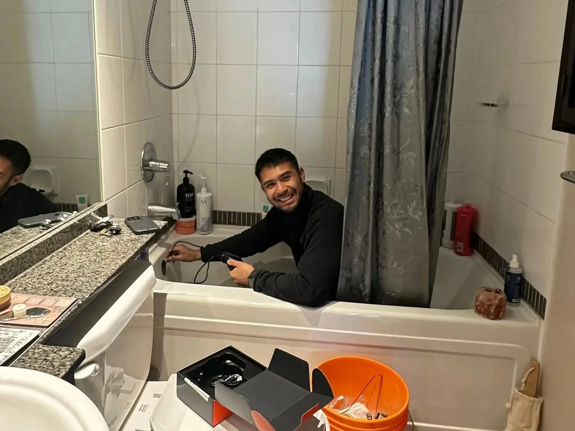 A man with a beard smiling in a bathtub while holding a hairdryer.