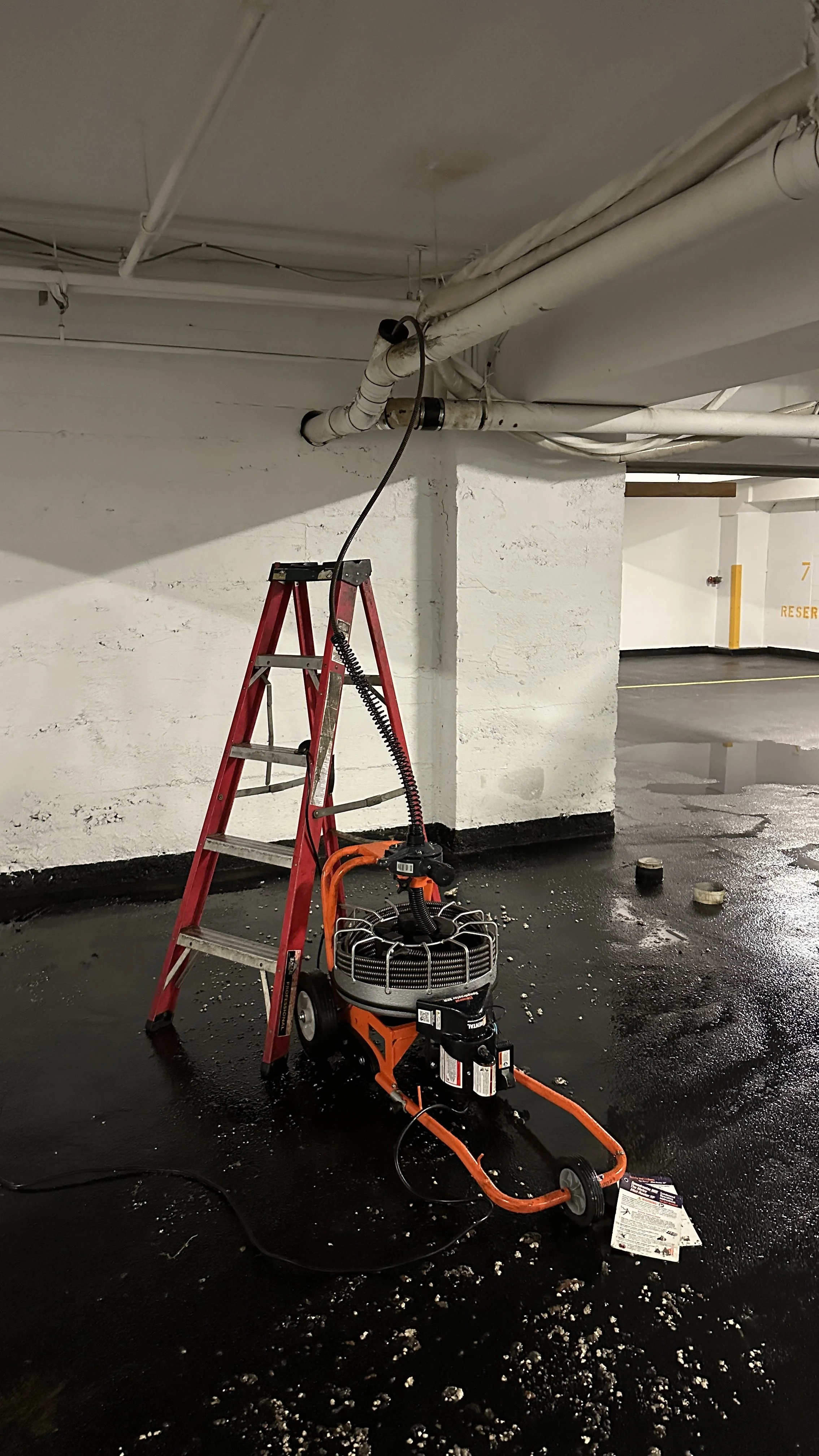 Pneumatic grinding machine on a wet parking garage floor, with a red step ladder nearby and pipes on the ceiling.